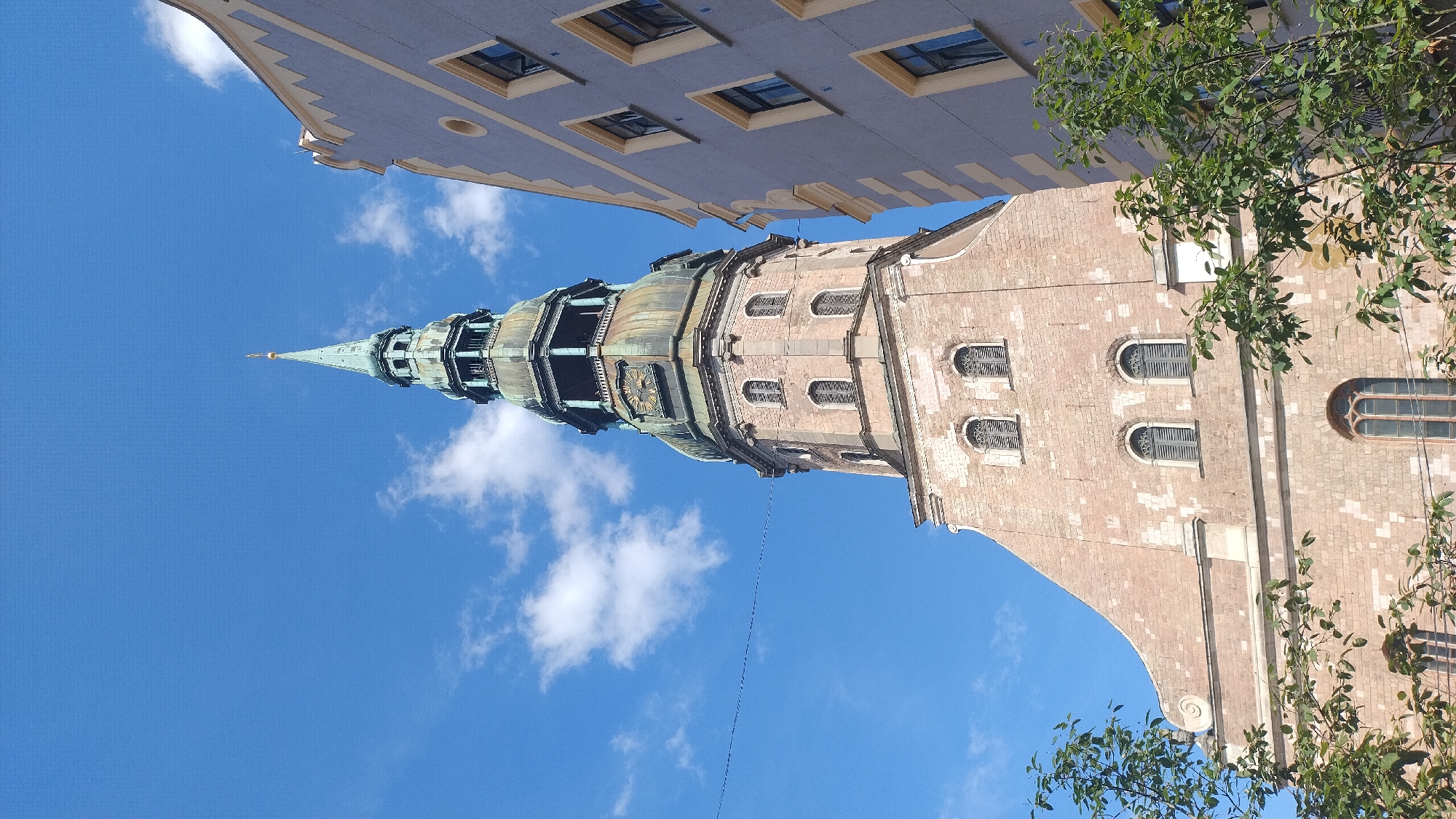 A view looking up at the spire of St. Peter's Church in Riga, Latvia, under a blue sky with scattered clouds. The brick tower of the church is tall and ornate, with a green copper spire at the very top, topped with a golden cross. The spire has multiple levels with windows and decorative elements. To the right of the church tower, a modern-looking building with a pale blue facade and cream-colored accents is visible, with several windows. Lush green trees with leaves frame the bottom and right side of the image. A thin wire stretches horizontally across the sky to the left of the church spire.