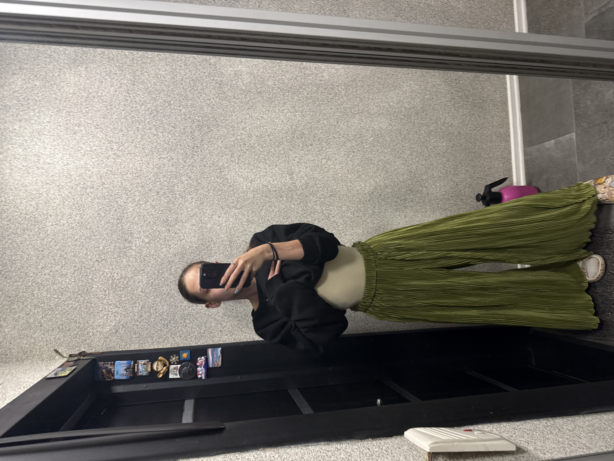 A young woman with her hair pulled back in a ponytail takes a mirror selfie in what appears to be a hallway. She is wearing a black sweatshirt, a beige or light-colored top underneath, and wide-leg green pleated pants. She is holding a black smartphone in her right hand, with her left hand touching her chest area. A pink spray bottle is visible on the floor behind her. The background consists of a textured wall, a black door with several magnets attached to it, and a full-length mirror. The floor is tiled. The scene is indoors, likely in a residential setting.