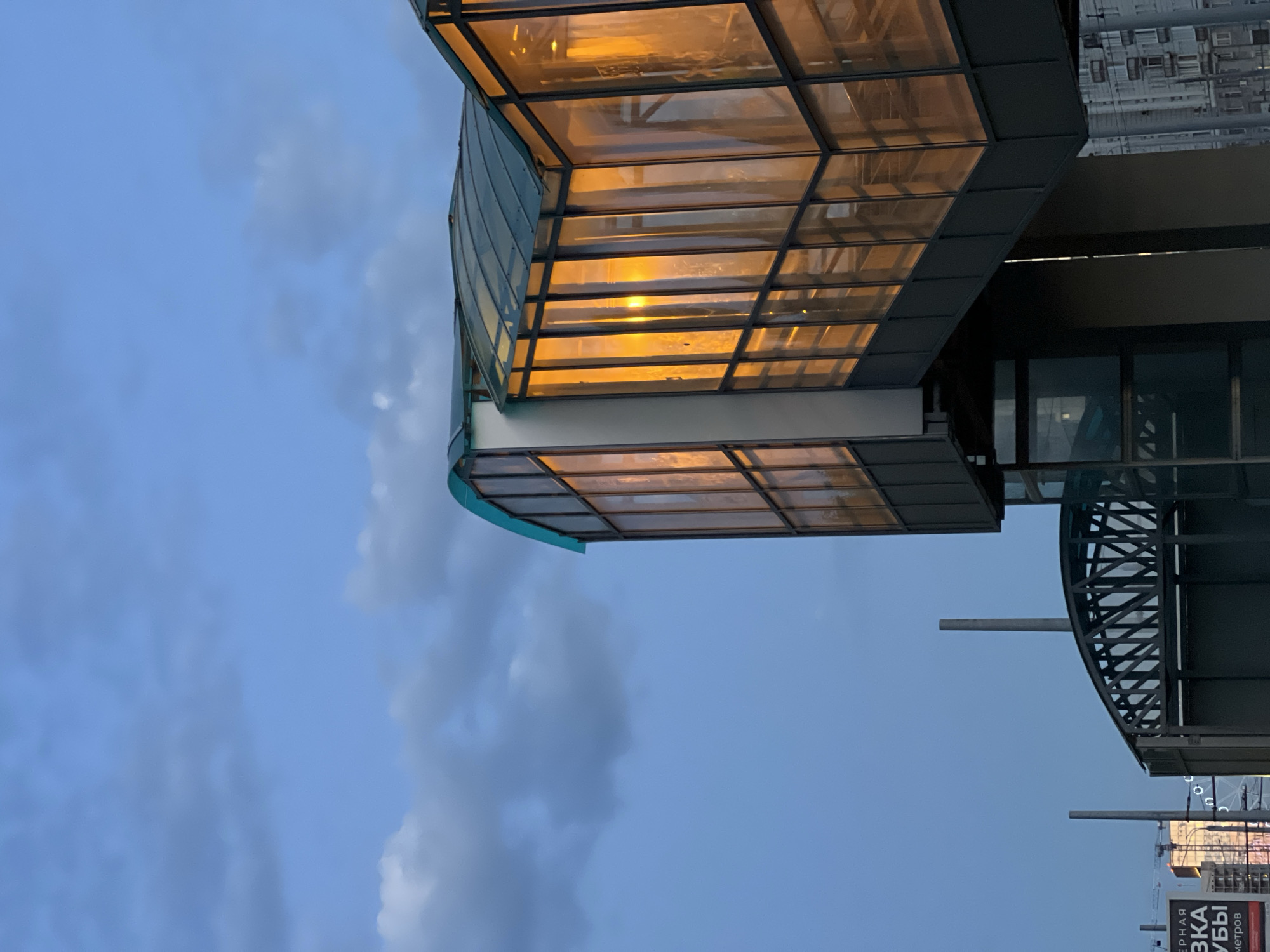 The image captures a contemporary pedestrian overpass in Kazan, Russia, at dusk. The overpass, constructed with a dark metal frame and large glass panels, is illuminated from within by a warm, orange light, suggesting interior lighting. The glass reflects the twilight sky and clouds, which are a muted blue with patches of darker gray clouds. The overpass has a distinctive curved roofline, composed of dark green material. Below the main overpass structure, another, more open section of the overpass with a metal lattice roof is visible. To the left, a tall, dark gray cylindrical structure, possibly a ventilation shaft or utility pole, rises from the ground. Further in the background, on the left side, a billboard with Cyrillic lettering is partially visible. The far background shows a city skyline with buildings and a Ferris wheel. The overall impression is one of urban infrastructure integrated into the evening cityscape. There are no people visible in the image.