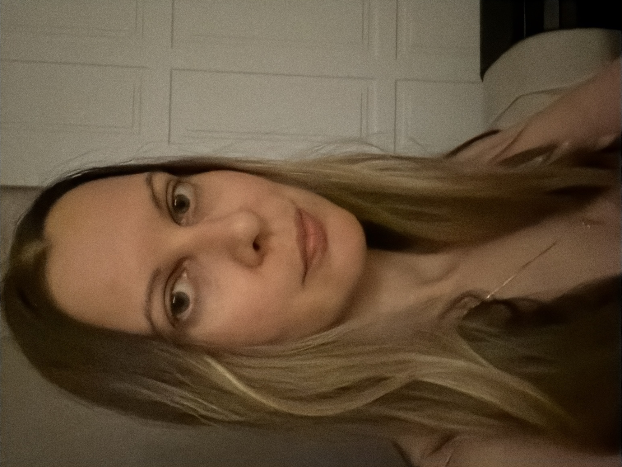 The image shows a headshot of a woman with long, blonde hair, looking directly at the camera. She has fair skin and appears to be indoors, with a plain, textured wall in the background. The lighting is soft and ambient. There are no discernible objects or actions that would indicate a specific location within the city of Izhevsk, Russia. The image is a close-up and does not provide any contextual clues about its surroundings.