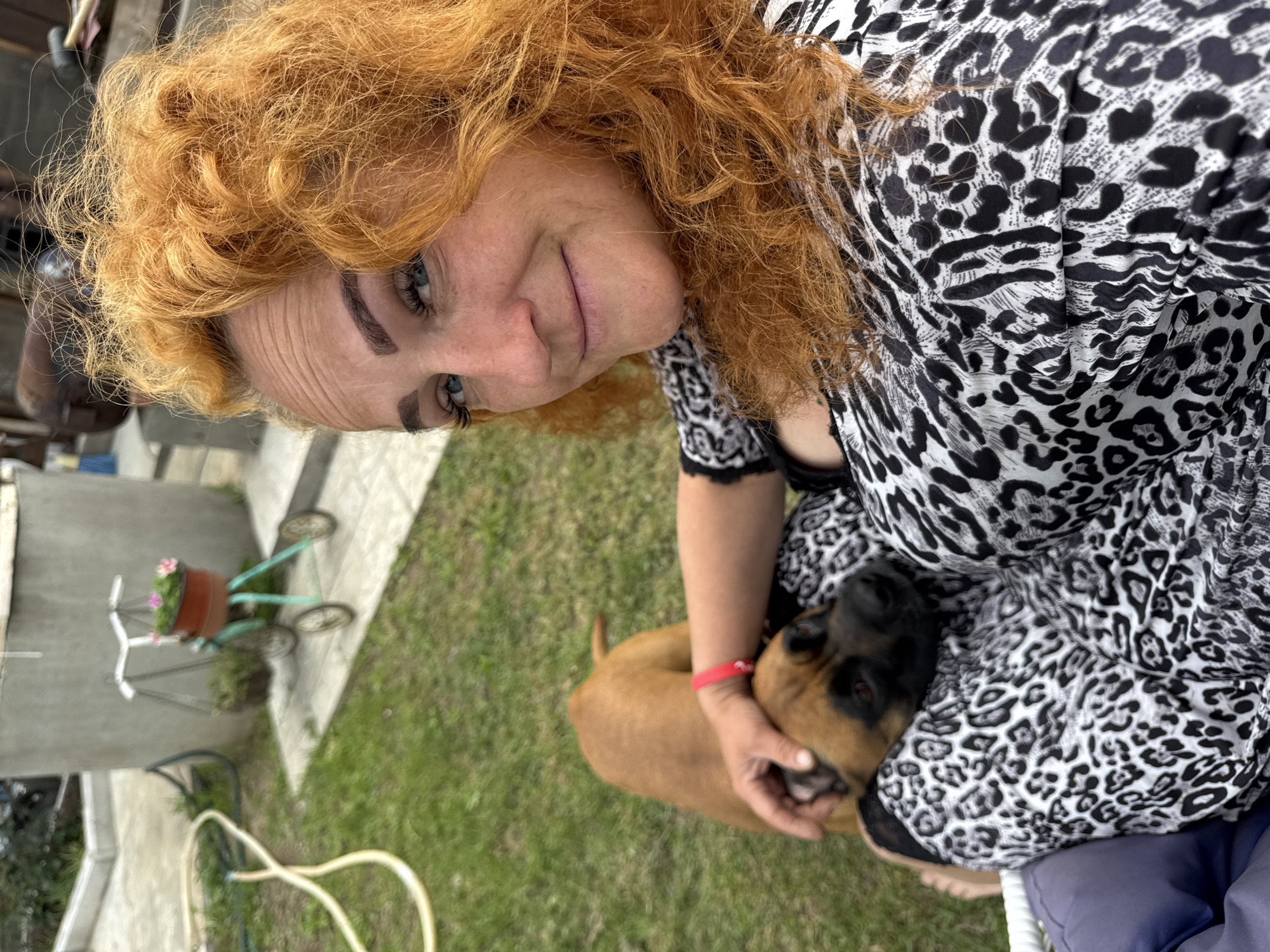 In an outdoor setting, a woman with vibrant red curly hair and striking blue eyes is captured in a close-up, selfie-style photograph. She is wearing a black and white leopard-print top. A young brown and black dog rests on her lap, with one of its ears being gently petted by the woman's hand, which is adorned with a red bracelet. The background reveals a grassy area with a paved pathway, a concrete cylindrical structure, and a small, decorative bicycle with planters. The overall scene suggests a relaxed, informal moment in a residential or garden area, potentially in the city of Chaikovsky, Russia, given the context provided.