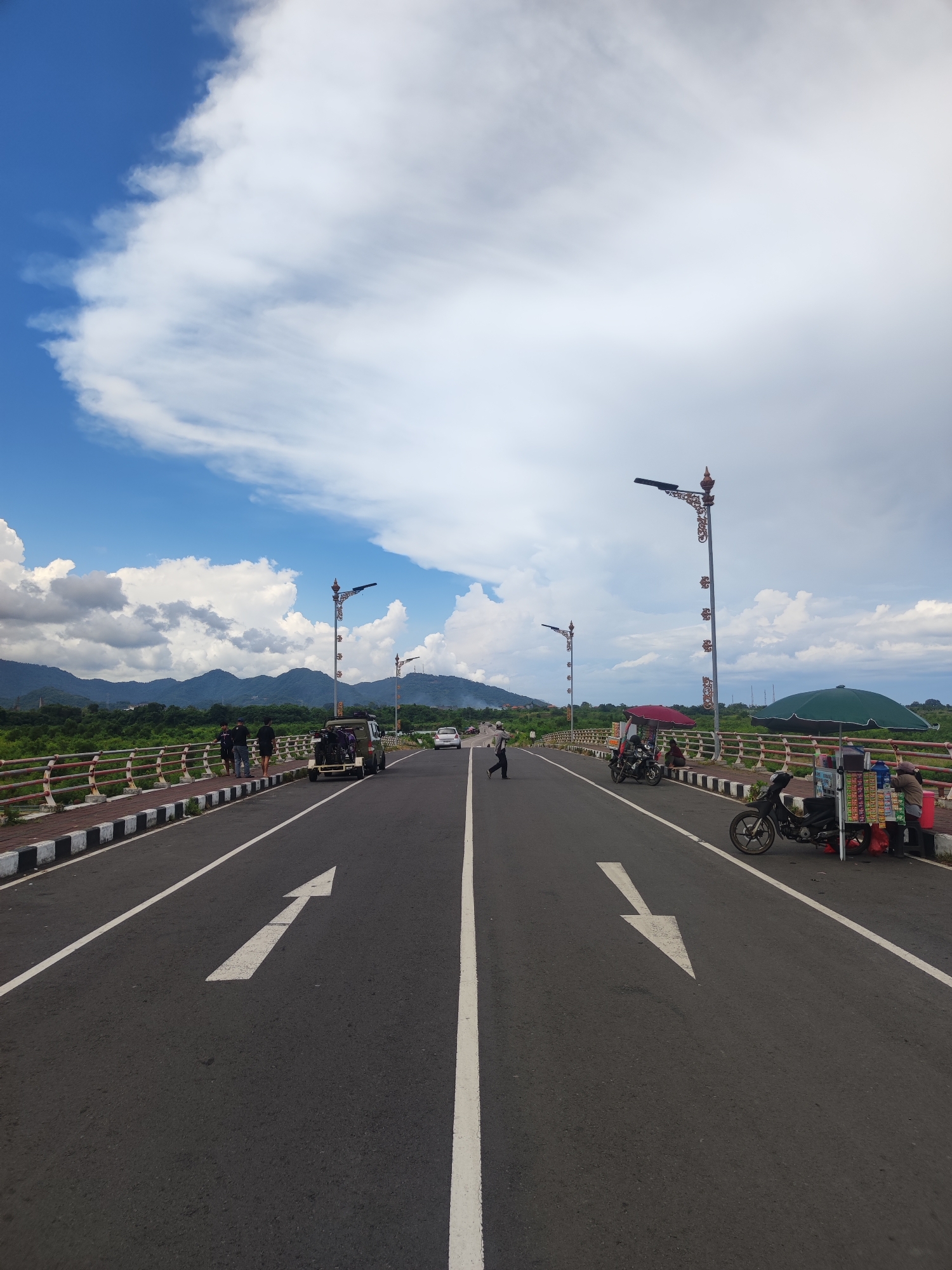 The image depicts a bridge in Jumpai, Indonesia, under a partly cloudy sky. A two-lane road with white arrows painted on the asphalt runs the length of the bridge. Along the sides of the road, black-and-white striped barriers border the bridge's railings, which are painted in a reddish-orange hue. Decorative streetlights are spaced along the bridge. On the left side of the road, several individuals are walking, and a vehicle is parked. On the right side, there are parked motorbikes and a makeshift stall selling snacks, with a person standing behind it. In the background, mountains are visible beneath the clouds. A person is seen crossing the road in the middle ground.