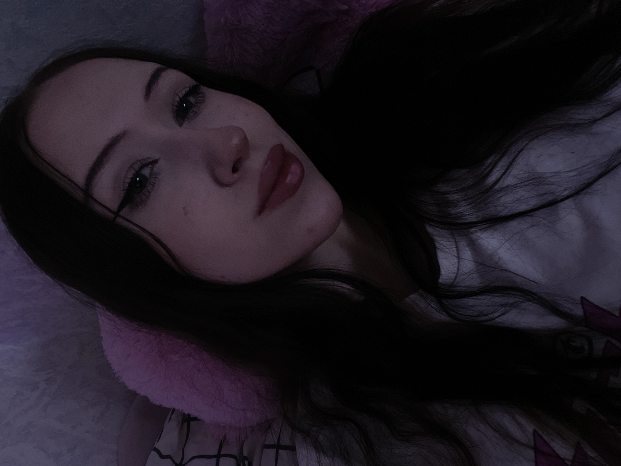 The image is a close-up, low-angle selfie of a young woman with long, dark hair, taken indoors. She is lying down, with her head resting on a soft, pink object that appears to be a pillow or stuffed animal. The background is dimly lit and appears to be a wall with some texture. The woman is wearing a white t-shirt. Her makeup includes winged eyeliner and lip gloss. The lighting is subdued, casting a purplish hue over the scene. There are no visible clues in the image that would allow for the identification of her location in Votkinsk, Russia, or any other specific city.