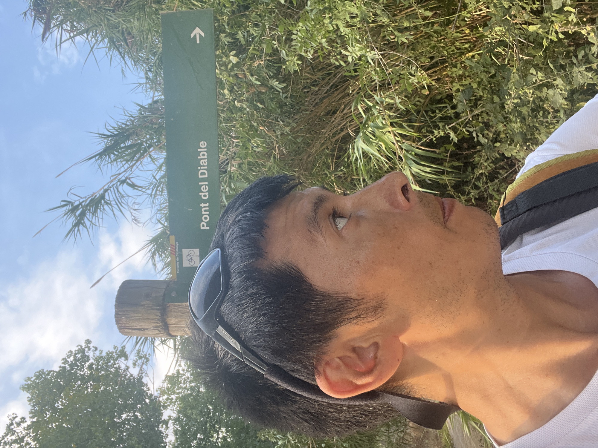 The image shows a man from the chest up, facing to the right, with sunglasses perched on his head. He is wearing a white t-shirt and has a backpack on. Behind him, a green sign with white text reads "Pont del Diable" and has an arrow pointing to the right. The sign also features a bicycling symbol and the text "Turisme de Catalunya". The background is lush with green foliage and tall grasses, under a bright blue sky with scattered clouds. This suggests a location within the natural surroundings of Tarragona, Spain, likely near a path or trail indicated by the sign. The sign itself refers to the Pont del Diable, a well-known Roman aqueduct located on the outskirts of Tarragona.