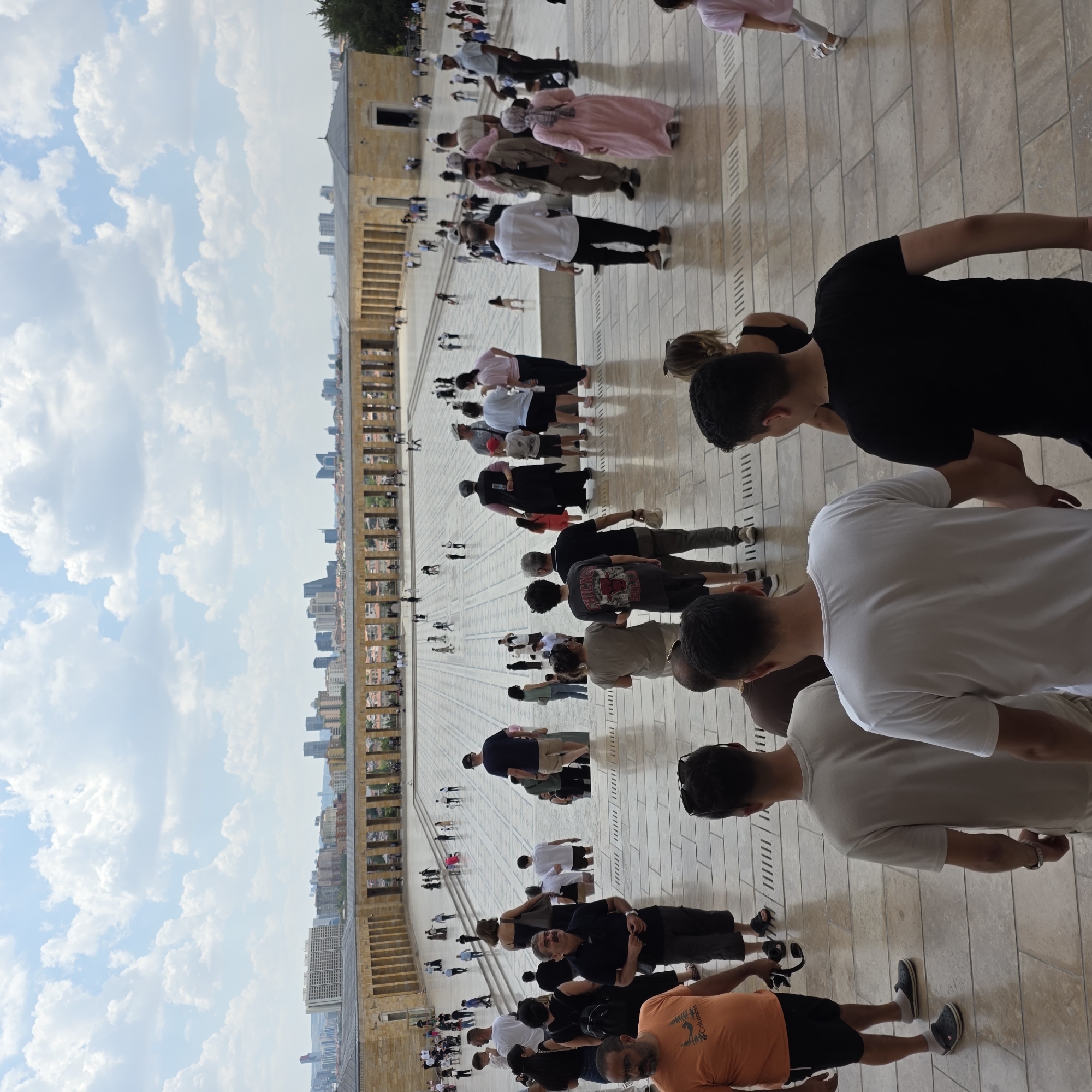 The image shows a view from a stone staircase looking out onto a large, open paved plaza in front of a long, colonnaded building. The plaza is filled with many people walking around. In the background, a city skyline with numerous tall buildings is visible under a partly cloudy sky. The people in the foreground are walking up or down the stairs, some are looking at the plaza, and others are facing away from the camera. The building appears to be a monument or memorial, with its classic architecture featuring stone columns and a wide entrance. The overall scene suggests a popular tourist destination on a clear day.