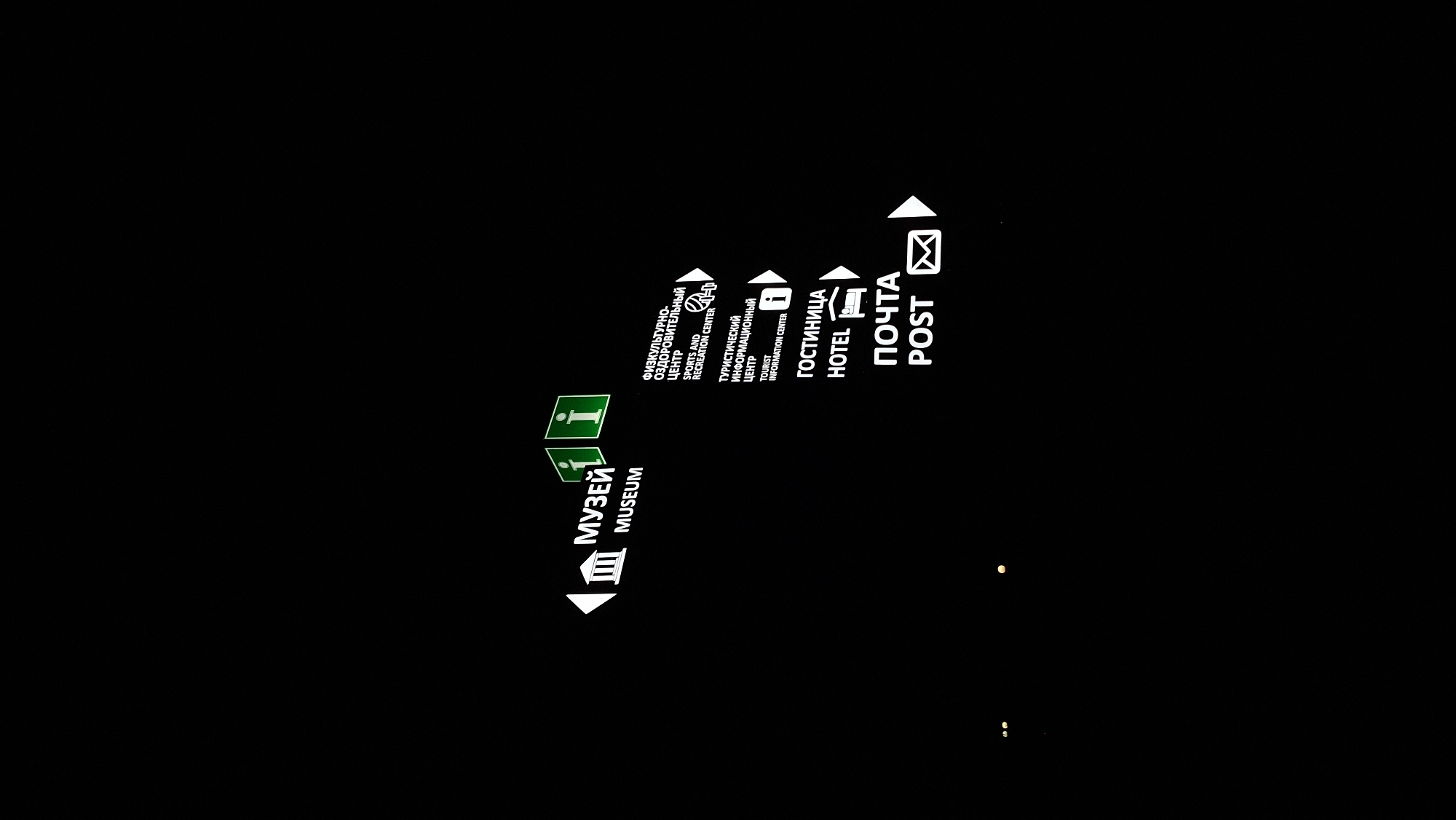 A night scene in Myadzyel, Belarus, features illuminated directional signs against a black background. The signs are white and green, indicating various locations.
To the left, a sign points to the left with the Russian word "МУЗЕЙ" (MUSEUM) and its English translation "MUSEUM" below, accompanied by an icon of a building with columns. Next to this, two green signs with a white "i" symbol for information are also visible.
To the right, a series of signs are stacked vertically. The topmost sign reads "ФИЗКУЛЬТУРНО-ОЗДОРОВИТЕЛЬНЫЙ ЦЕНТР" (SPORTS AND RECREATION CENTER) with an icon resembling a person playing basketball. Below this is a sign for "ТУРИСТИЧЕСКИЙ ИНФОРМАЦИОННЫЙ ЦЕНТР" (TOURIST INFORMATION CENTER) with a white "i" icon, pointing to the right. Further down, a sign for "ГОСТИНИЦА" (HOTEL) with a bed icon points to the right. The bottommost sign reads "ПОЧТА" (POST) with an envelope icon, also pointing to the right.
The overall environment appears to be a dark outdoor setting, likely a street or public area at night, with only the signs being illuminated.