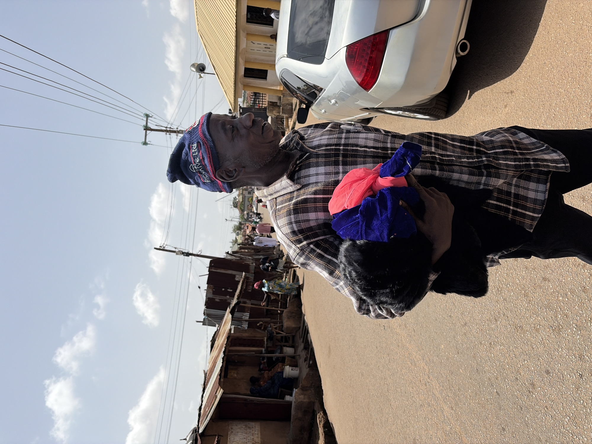 An elderly, dark-skinned man wearing a blue and red knit cap, a brown and white plaid shirt, and black pants stands on a paved street holding a bundle of colorful cloths. He is looking to the left. A white car is parked nearby. Across the street are dilapidated buildings with corrugated metal roofs. A few people are visible in and around the buildings. Utility poles and wires stretch across the sky. The sky is pale blue with scattered clouds. This appears to be a street scene in Jos, Nigeria.