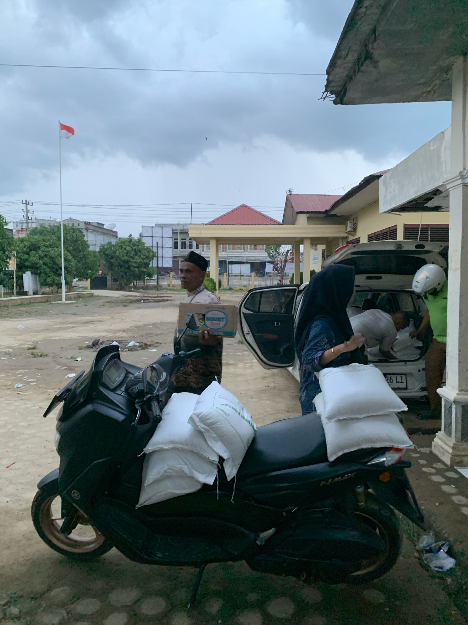 In Pantegurah, Indonesia, under a cloudy sky, a black motorcycle is parked on a dirt lot in front of a building. The motorcycle is laden with several white sacks, two of which are prominently stacked on the seat. A man in a light-colored shirt and black cap stands near the motorcycle, holding a cardboard box labeled "MOUNT DRINK." In the background, a white car with its trunk open is being loaded with sacks by at least two individuals. One person, wearing a hijab, stands beside the car. Another person wearing a green shirt and white helmet is seen inside the trunk. An Indonesian flag flies on a pole in the distance. The environment appears to be a public space, possibly near a government building or community center, with some debris scattered on the ground.