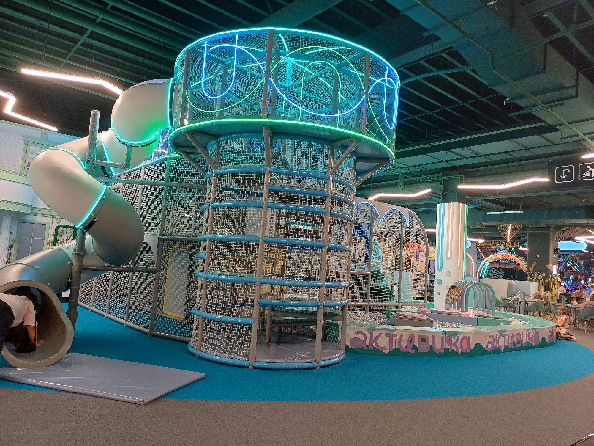 An indoor playground with a large, multi-level play structure dominates the scene. The structure features a prominent cylindrical tower with a blue net enclosure and vibrant neon blue and green spiral lights. Attached to the tower is a large, curved, pale gray slide, also outlined with neon blue lights. A person in a light pink shirt and dark pants is partially visible entering the mouth of the slide. The playground is situated on a bright turquoise carpet. In the background, more play structures and seating areas are visible. The ceiling is dark with exposed beams and industrial lighting, including some horizontal orange neon lights. There are also signs on the wall in the background, one showing icons of a downward arrow, a wheelchair, and a person with a suitcase. The text "активка" is written in large, colorful letters along the edge of a ball pit area in the middle ground. The overall impression is a modern, well-lit, and engaging indoor entertainment space.