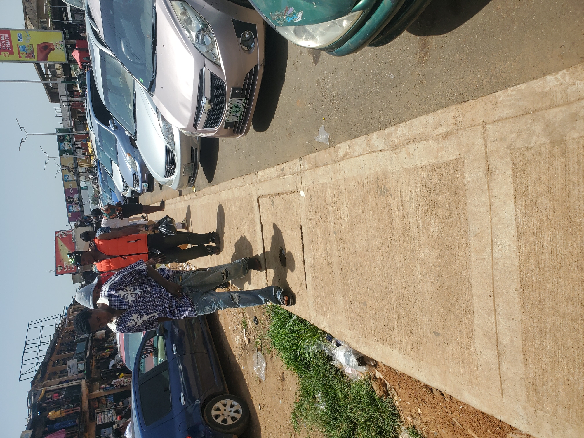 The image depicts a street scene in Jos, Nigeria, on a sunny day. A sidewalk made of concrete is visible in the foreground, with some dirt and patchy grass along its edge. A man wearing a checkered shirt, ripped jeans, and sandals is walking on the sidewalk. Behind him, another man in an orange vest and a third person in a grey hoodie are also walking. Several cars are parked along the street, including a blue car on the left and several silver and light pink cars on the right. The background shows a row of buildings with shops and stalls, and overhead billboards advertising various products. The overall impression is of a busy urban environment.