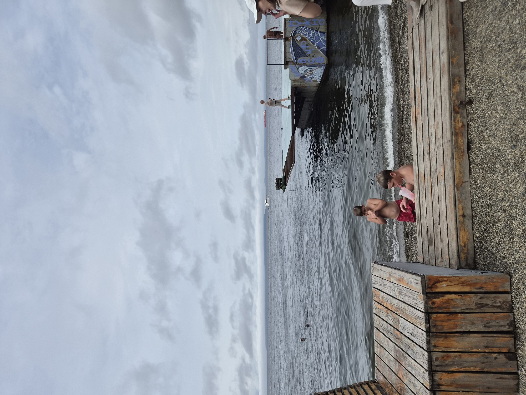 The image depicts an outdoor scene by the sea, likely in Vladivostok, Russia, given the presence of a body of water and a pier. In the foreground, there is a wooden structure with a slatted surface, appearing to be a bench or seating area. Adjacent to it is a pebble beach.
Several individuals are present in the image. Two young boys are on the beach near the water's edge. One boy, wearing red shorts, is standing and appears to be holding his hands to his chest. Another boy is sitting and holding a pink cup. Further in the water, two individuals are visible as small dark shapes, suggesting they are swimming.
In the background, a pier extends into the sea. On the pier, a man in light-colored clothing is walking, and another man, who is shirtless, stands near the edge. A person in the far right of the frame, wearing a white cap, is partially visible. There are boats and a large ship in the distance on the water. The sky is overcast with clouds. The overall atmosphere suggests a cool, possibly summer day at a coastal location.