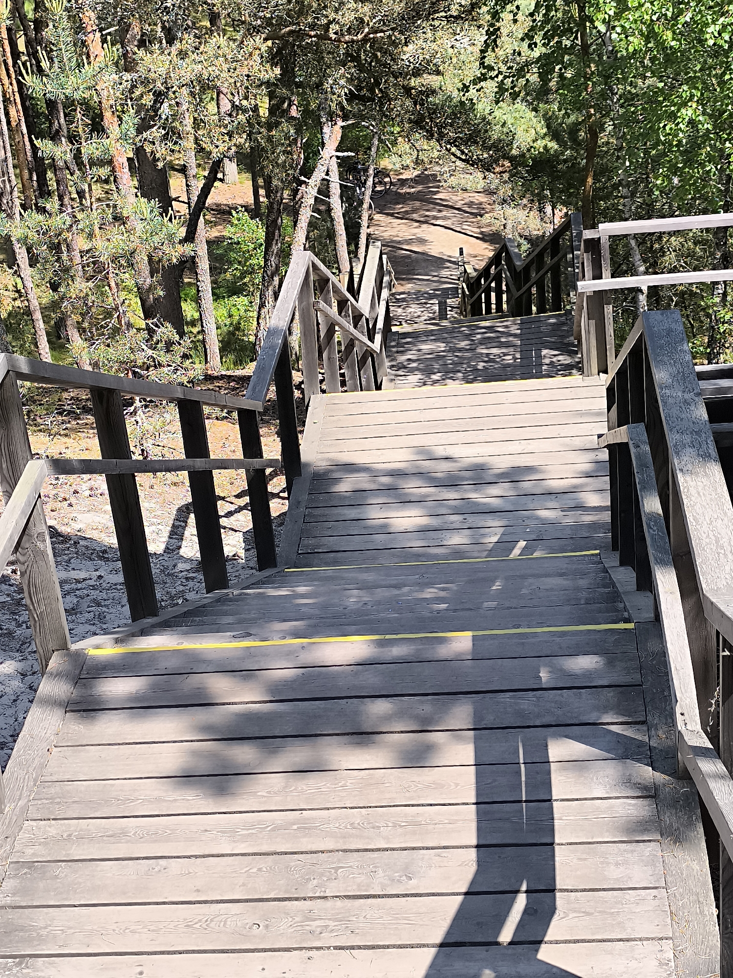 The image shows a wooden staircase descending through a forest in Neringa, Lithuania. The staircase is constructed with wooden planks and has a wooden railing on both sides. Yellow tape has been applied to some of the steps, likely as a safety measure. The surrounding environment is a dense forest with numerous trees, primarily pines, casting shadows on the stairs. The ground beneath the stairs is sandy, typical of the Curonian Spit's dunes. The lighting suggests it is daytime, with sunlight filtering through the tree canopy. In the distance, at the bottom of the stairs, a sandy path is visible, along with what appears to be a bicycle.