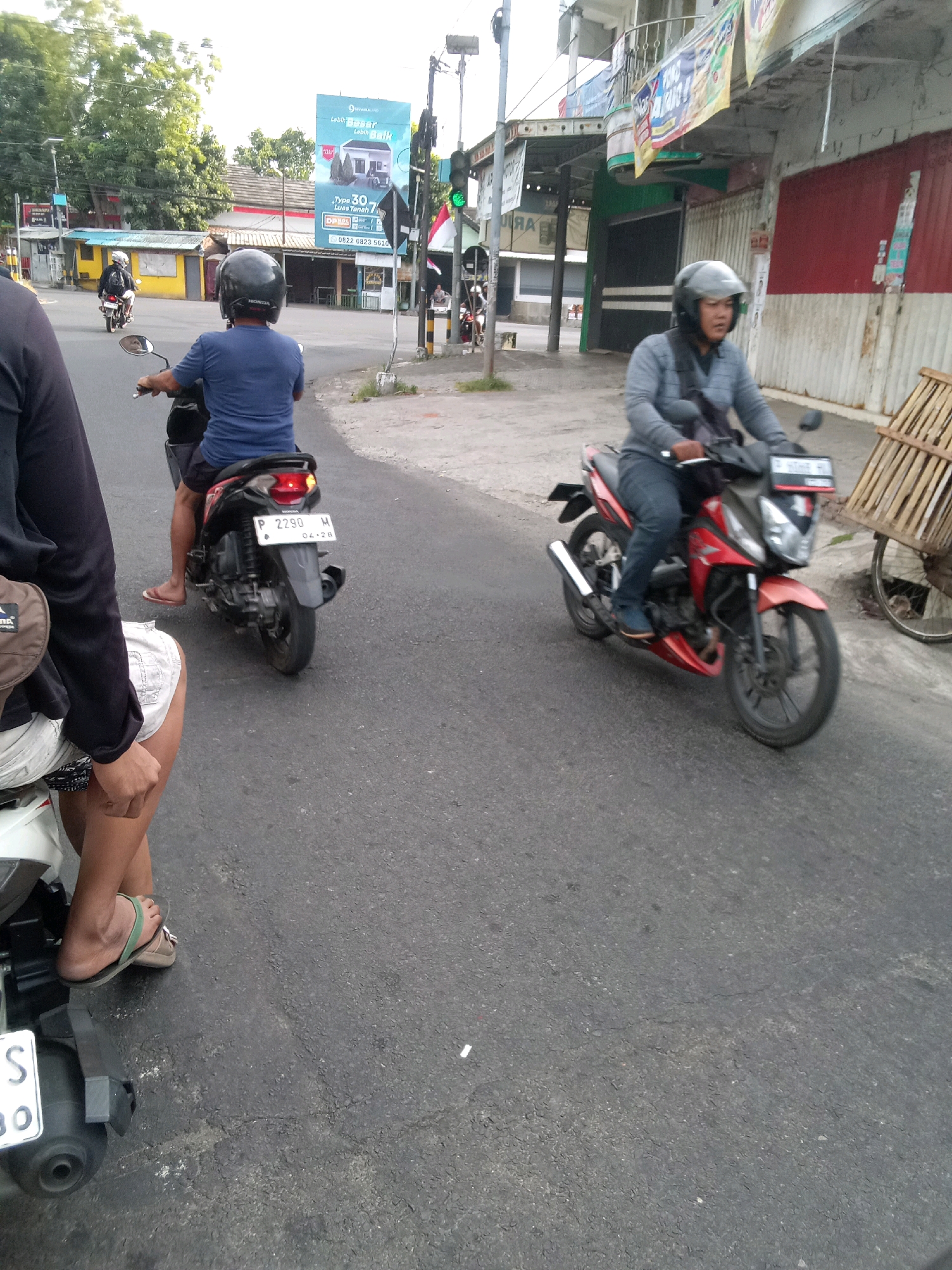 The image depicts a street scene in Jember, Indonesia, focusing on motorcycle activity. In the foreground, a person wearing a black long-sleeved shirt and white shorts is seated on a white scooter, their legs visible. To the left, another scooter's license plate is partially visible, reading "S 80". In the middle ground, a person in a blue shirt and dark shorts rides a black motorcycle, wearing a black helmet. The motorcycle has a license plate that reads "P 2290 H 04.26". Further ahead, on the right side of the image, a person in a grey long-sleeved shirt and blue jeans rides a red motorcycle, also wearing a grey helmet. This motorcycle has a license plate with what appears to be Indonesian script. The background shows buildings, trees, and a billboard advertising housing. The street is paved with asphalt. Traffic lights are visible on a pole in the middle-left of the image. The overall impression is a typical street environment with ongoing motorcycle traffic.