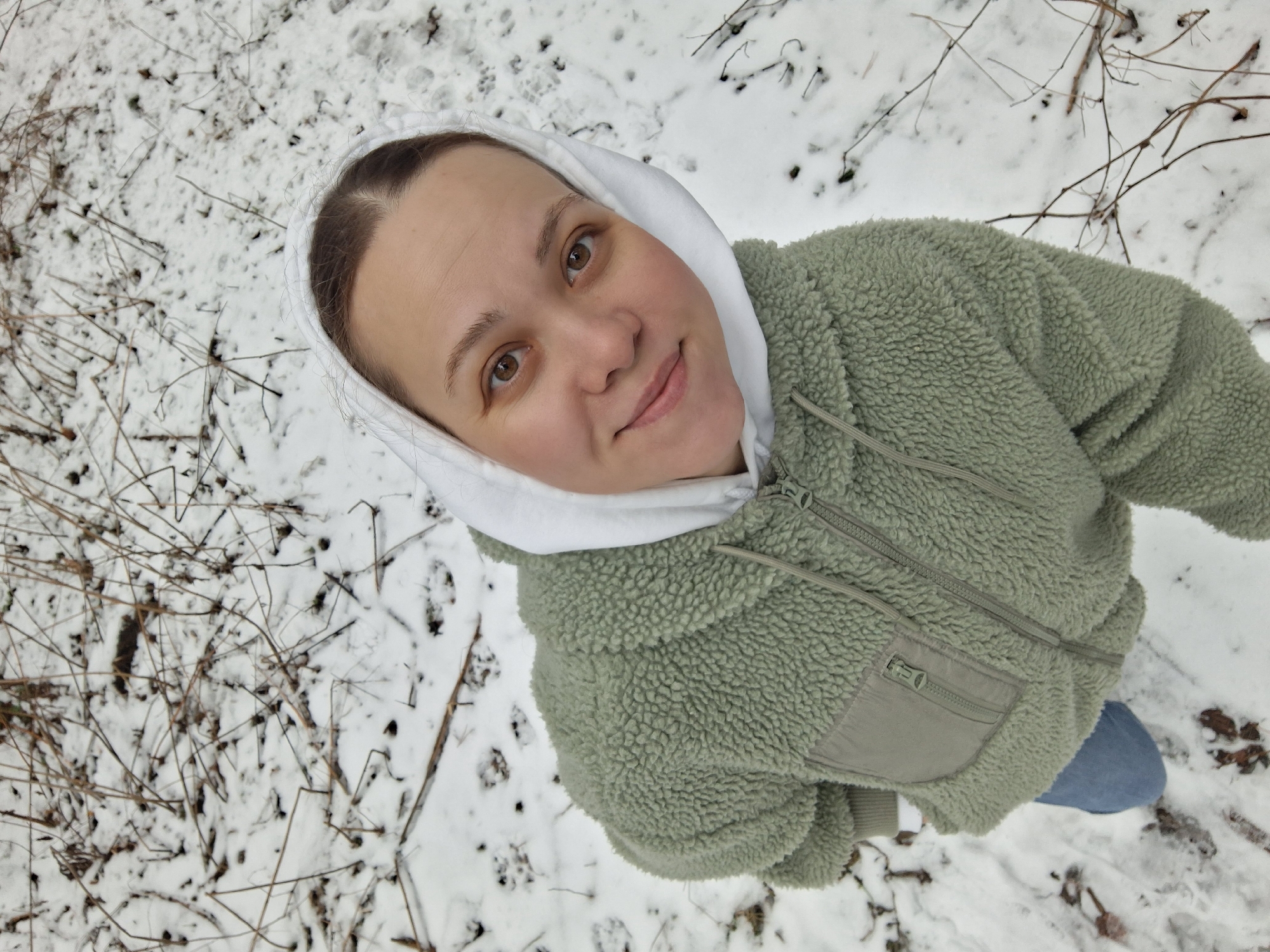 A woman stands in a snowy area, likely a park or wooded area, as indicated by the snow-covered ground and scattered twigs. The woman is wearing a fuzzy, sage green jacket with a white hood. She is looking directly into the camera with a slight smile. Her face is visible, showing her eyes, nose, and mouth clearly. A patch of blue denim, likely jeans, is visible at the bottom edge of the frame. The background shows a ground covered in snow mixed with scattered dry branches and twigs. The photo appears to have been taken from a high angle, looking down at the woman. No specific landmarks are identifiable to confirm the city of Chaikovsky, Russia.
