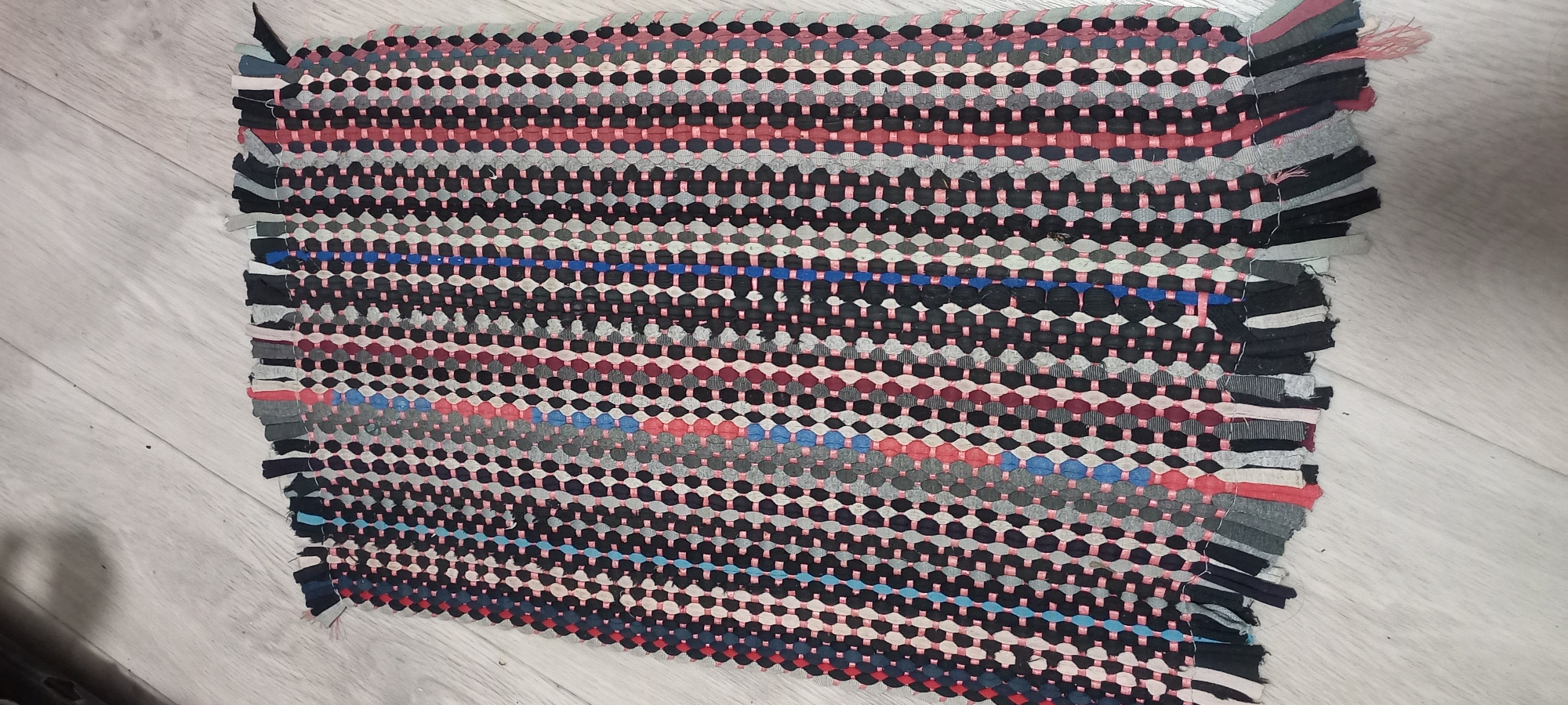 A close-up, slightly angled shot shows a handmade rag rug lying on a light-colored wooden floor. The rug is rectangular and oriented horizontally, with its top edge facing away from the camera. The rug is constructed from strips of fabric woven together, creating a textured surface with a pattern of alternating dark and light oval shapes. Vertical stripes of various colors, including black, gray, pink, and blue, run the length of the rug. The bottom edge of the rug is unfinished, with frayed fabric strips hanging down, resembling fringe. The floorboards are visible beneath the rug, running horizontally across the frame. The lighting appears to be natural or overhead, casting soft shadows. No people or animals are present in the image. The setting is indoors, on a floor. The location is specified as Идрисово, Russia.
