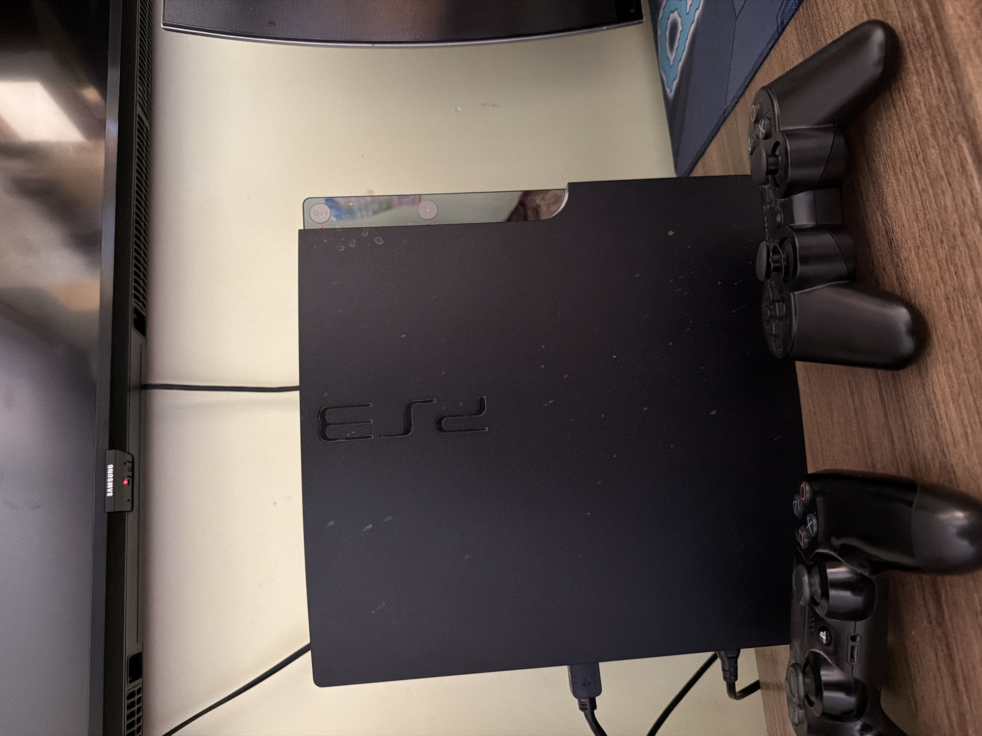 The image shows a black Sony PlayStation 3 console placed vertically on a wooden surface. Two black PlayStation controllers are positioned in front of the console. The console has the "PS3" logo embossed on its side. A cable is connected to the front of the console. In the background, a portion of a Samsung television is visible, with a red indicator light on. The environment appears to be an indoor setting, likely a room, with a plain wall behind the console. The overall impression is a still-life arrangement of gaming equipment. There is no information or visual cues to suggest the location is in Samarkand City, Uzbekistan.
