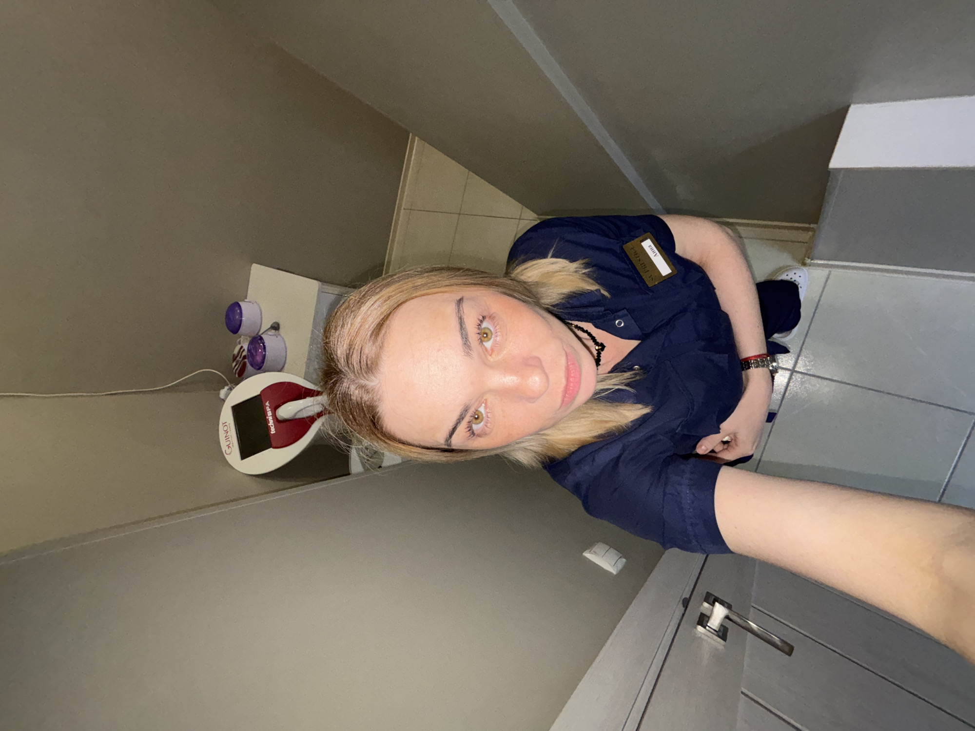 A selfie taken from a low angle, showing a person with blonde hair and hazel eyes, wearing a dark blue uniform with a name tag that reads "Anna", positioned inside a room with a tile floor. The person is wearing a black choker and a watch on their left wrist. Visible to the left of the person, on a white surface, is a piece of equipment with purple details and a "GUINOT" labeled device. The room has neutral-toned walls and is in Unknown, Russia.