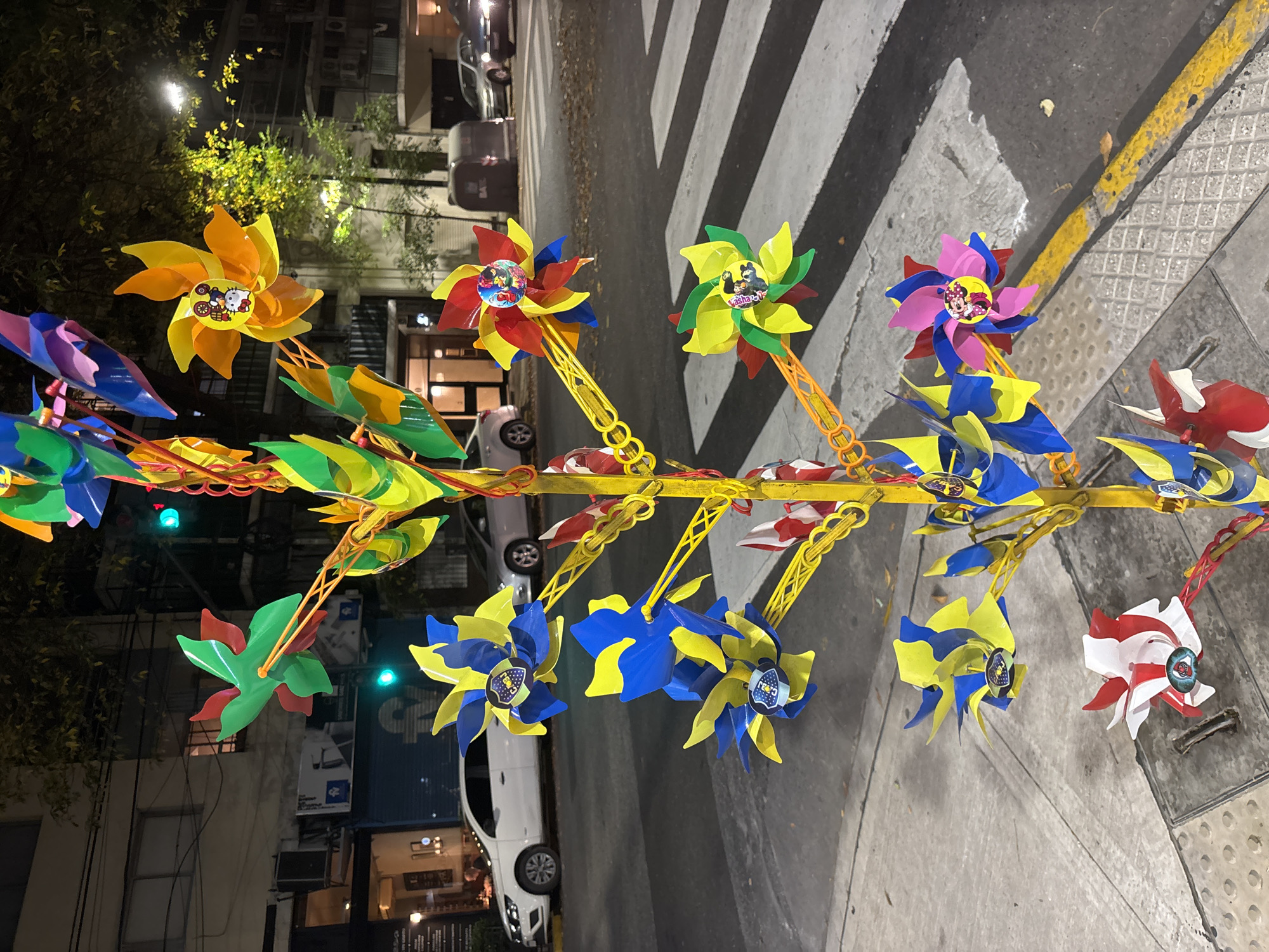 The image shows a street display of colorful pinwheels with cartoon character stickers in Buenos Aires, Argentina. The pinwheels are attached to a yellow metal frame and arranged on the sidewalk, next to a crosswalk. In the background, there are buildings, trees, and parked cars. The scene appears to be at night, with streetlights illuminating the area.