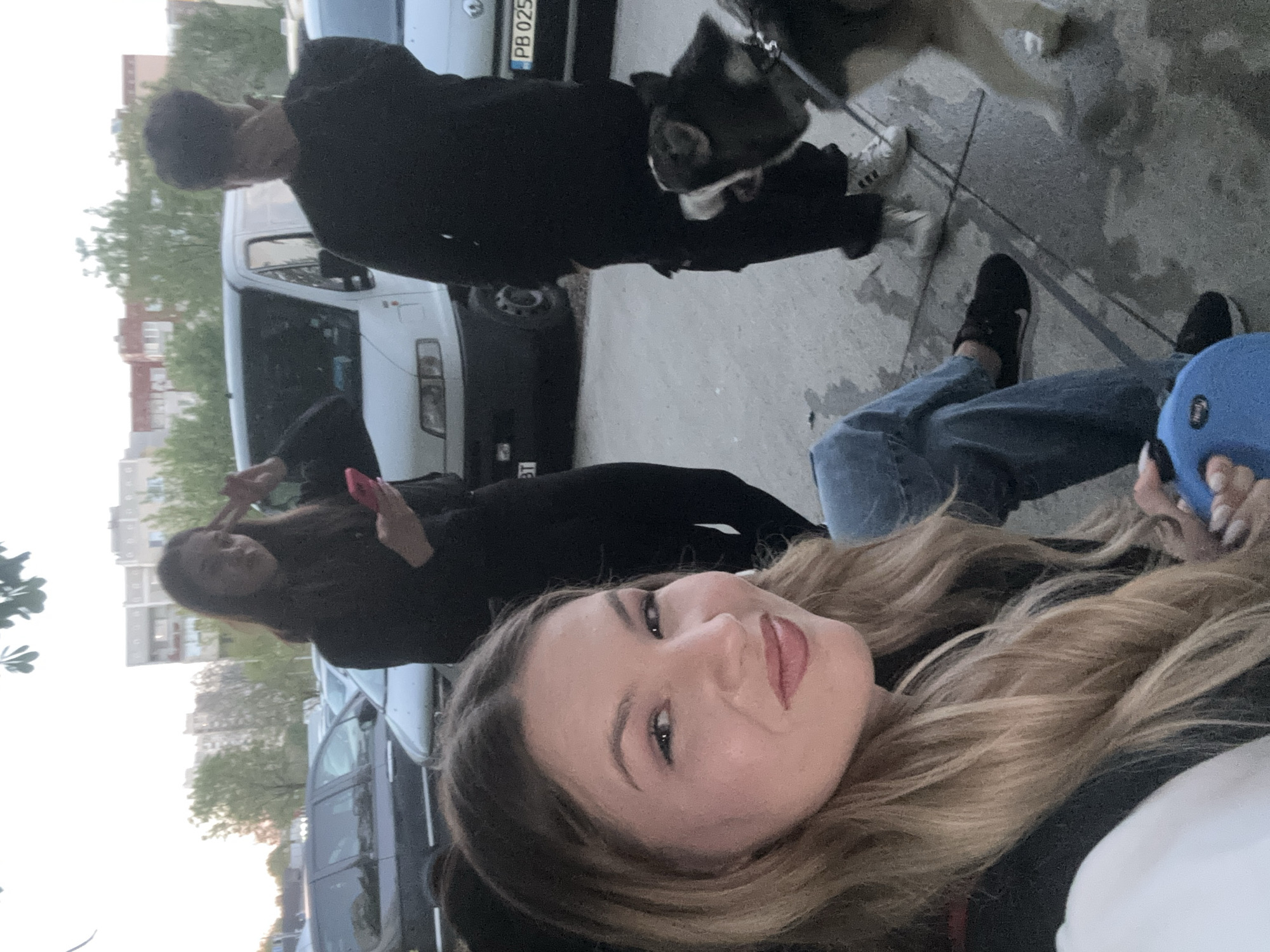 The image shows a selfie-style photograph taken outdoors. A woman with long blonde hair is positioned in the foreground. Behind her, another woman is holding up two fingers in a peace sign and holding a red cell phone. To the left, the background includes a partially visible row of parked cars and some buildings in the distance. To the right a partially visible person wearing blue jeans, a white shirt, and a blue hat. Behind the first woman in the foreground a man is walking a black and white dog.
In the background, a white van is parked, with a license plate number PB 0253 IT visible. The location appears to be an urban setting. I am unable to confirm the location in Plovdiv, Bulgaria.