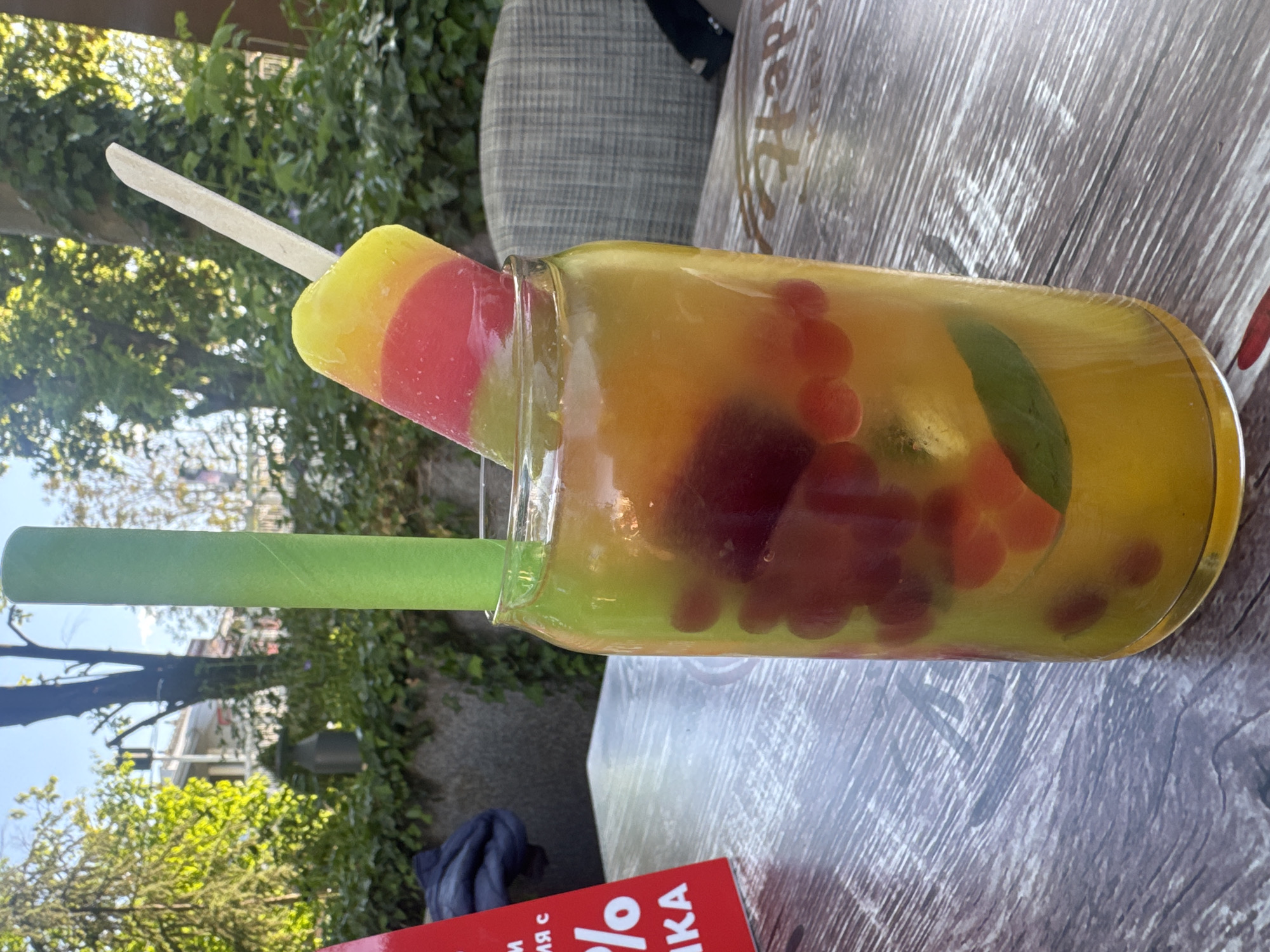 A medium shot depicts a beverage in a glass jar on a weathered grey wooden surface, outdoors in what appears to be a café or similar setting. The beverage contains dark red fruit or berries and possibly mint leaves suspended in a yellowish liquid. A popsicle with a layered yellow, red, and green color scheme is placed in the mouth of the jar, along with a wide green straw. Vegetation is visible in the background, including what looks like trees and shrubbery. The edge of a red and white printed sign is visible in the lower foreground. The overall impression is that of a refreshing drink served outdoors on a sunny day. The location in the city of Plovdiv, Bulgaria cannot be determined.