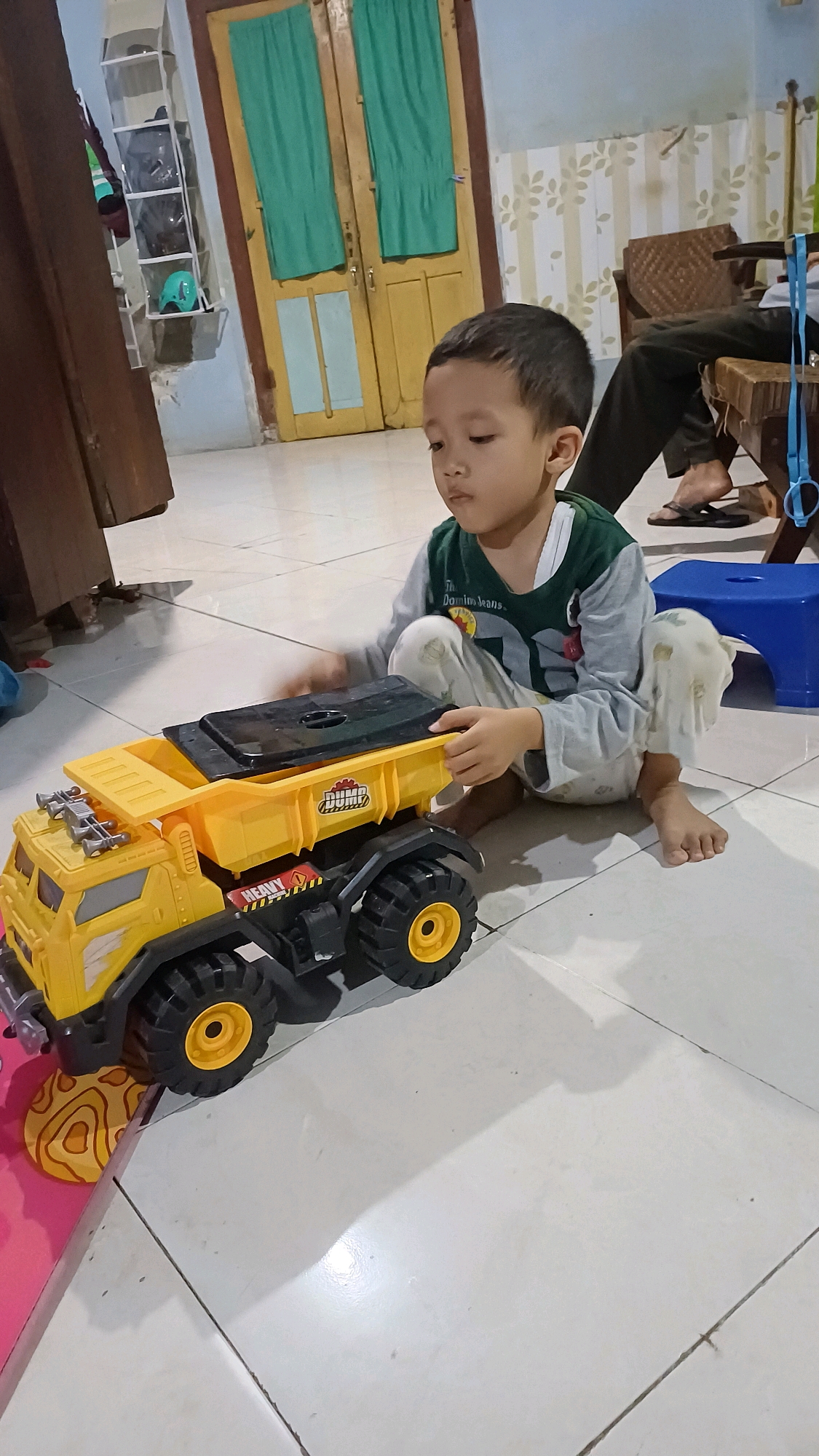 A young East Asian boy is crouching on a tiled floor, intently playing with a large yellow toy dump truck. He is wearing a grey long-sleeved shirt with a green collar and white accents, and light-colored pants that are draped around his legs. His left hand is on the truck's black covering, and his right hand is near the truck's bed. The dump truck is detailed with black tires, yellow rims, and "DUMP" and "HEAVY" text. To the left of the truck, a pink object with a yellow circular pattern is partially visible. In the background, a brown wooden door with two green curtains is visible, along with a white shelf unit containing various items and a blue plastic stool. The floor is made of off-white square tiles. An adult's legs, wearing dark pants and sandals, are visible in the upper right corner of the image. The lighting suggests an indoor environment, possibly a home in Wonokerso, Indonesia.