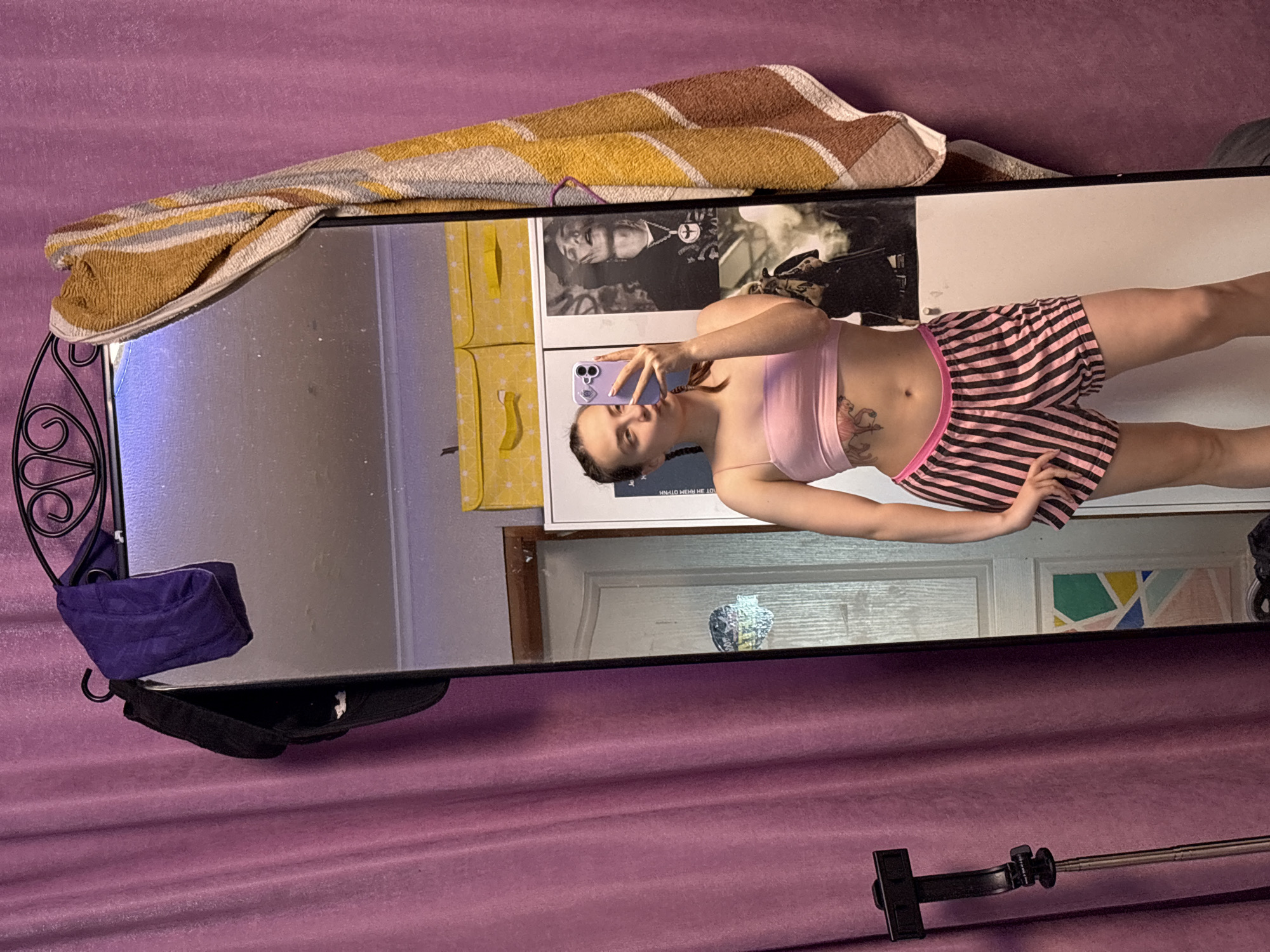 A young woman is depicted taking a mirror selfie in an indoor setting. She stands upright, holding a light purple smartphone with multiple cameras to capture her reflection in a large, full-length mirror. She is dressed in a light pink sports bra or crop top and pink and black striped shorts, revealing a tattoo on her left side/rib cage area.
The mirror's frame, adorned with decorative black metalwork on its left side, has a multi-colored towel (yellow, brown, white, and grey) draped over its top edge. A small purple pouch hangs from the top left of the frame, and a black cap rests on its bottom left.
Reflected in the mirror, behind the woman, are elements of the room. Two framed artworks are visible: a black and white poster on the left features a person, possibly a musician, with a chain, displaying partially legible text like "HN-LLO HE LOV". The poster to its right shows abstract geometric patterns in various colors. A yellow piece of furniture or shelving is reflected to the far left of the posters, and a white paneled door appears below them.
Outside the mirror's view, a vibrant purple fabric or curtain covers the wall on the right. A black and silver extendable tripod or stand is situated on the floor in the bottom right corner of the image. The scene, likely a personal space such as a bedroom in Прикамский, Russia, appears to be well-lit, suggesting daytime. The woman's expression is neutral and focused on taking the photo.