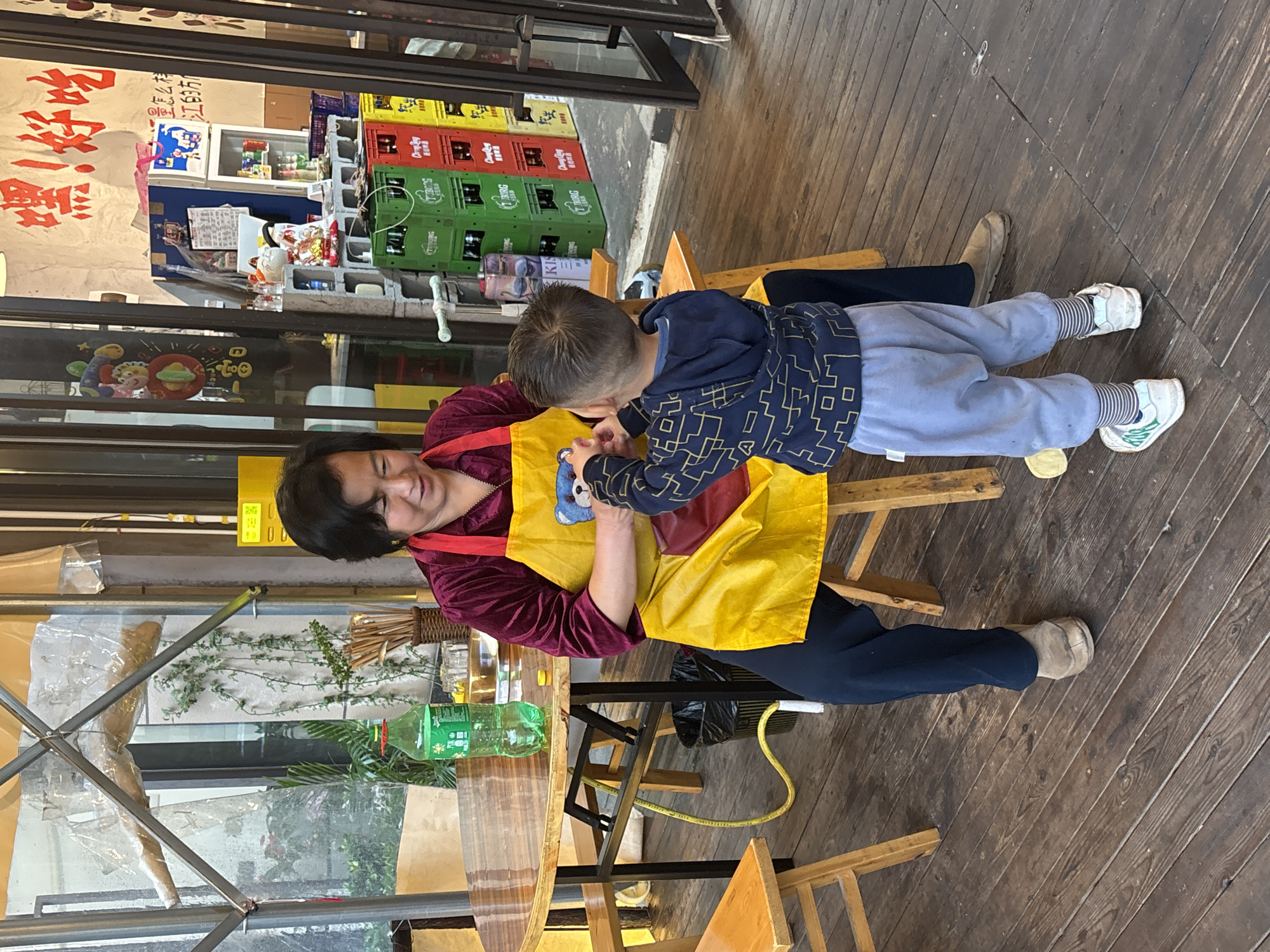 A woman wearing a maroon top and dark pants, with a yellow apron, is seated on a wooden chair and holding a young boy. The boy wears a blue geometric-patterned jacket and light blue pants. They appear to be inside or just outside a building with a wooden floor. A table with a green plastic bottle sits beside them, and additional wooden chairs are visible. Behind them, stacks of colorful crates and a sign with Chinese characters are visible. Based on the sign, this is possibly a restaurant located in the city of Jiangjin, China.