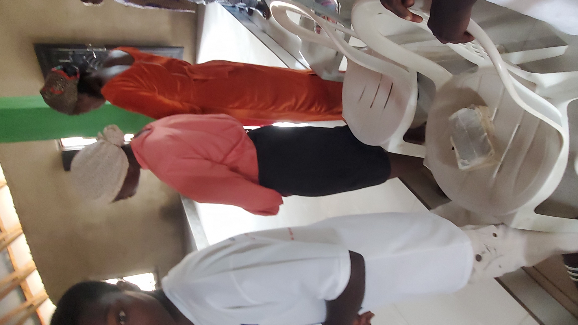 In an indoor setting, three individuals are visible, facing away from the camera. The person on the left is wearing a white t-shirt and light-colored pants. To their right, a person in a peach-colored shirt and black skirt is standing. Further to the right, another person is wearing an orange dress. Two white plastic chairs are present in the foreground, with a small white table between them. On the table, there is a rectangular object wrapped in clear plastic, possibly a pack of tissues or a small box. The environment appears to be a simple indoor space with a wooden ceiling structure and plain walls.