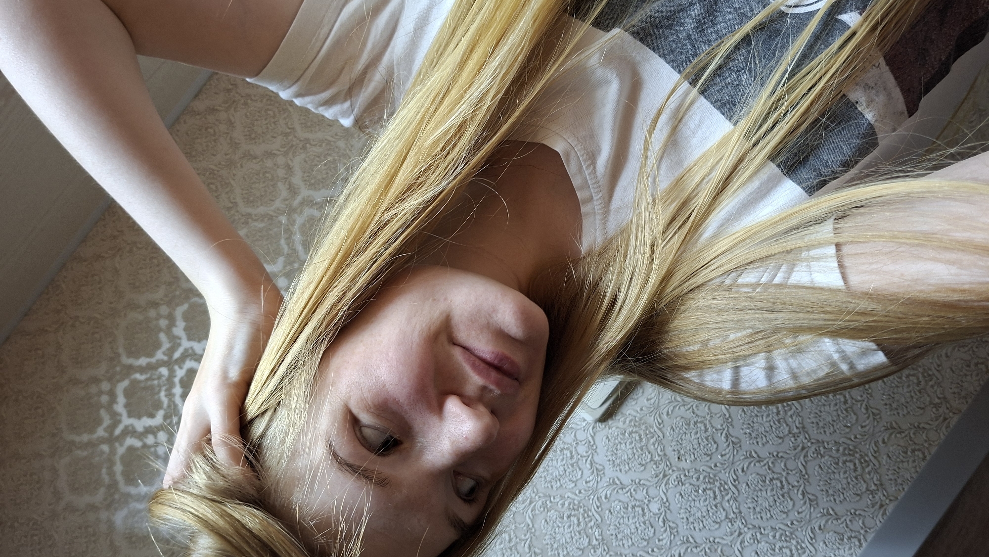 A young Caucasian woman with long blonde hair is shown in a selfie. Her left arm is bent and resting on her head, with her hand touching her hair. She is looking to her left with a slight smile. She is wearing a white t-shirt with a grey and black pattern. The background is a patterned wallpaper. The image appears to have been taken indoors in the city of Bolshoy Bukor, Russia.