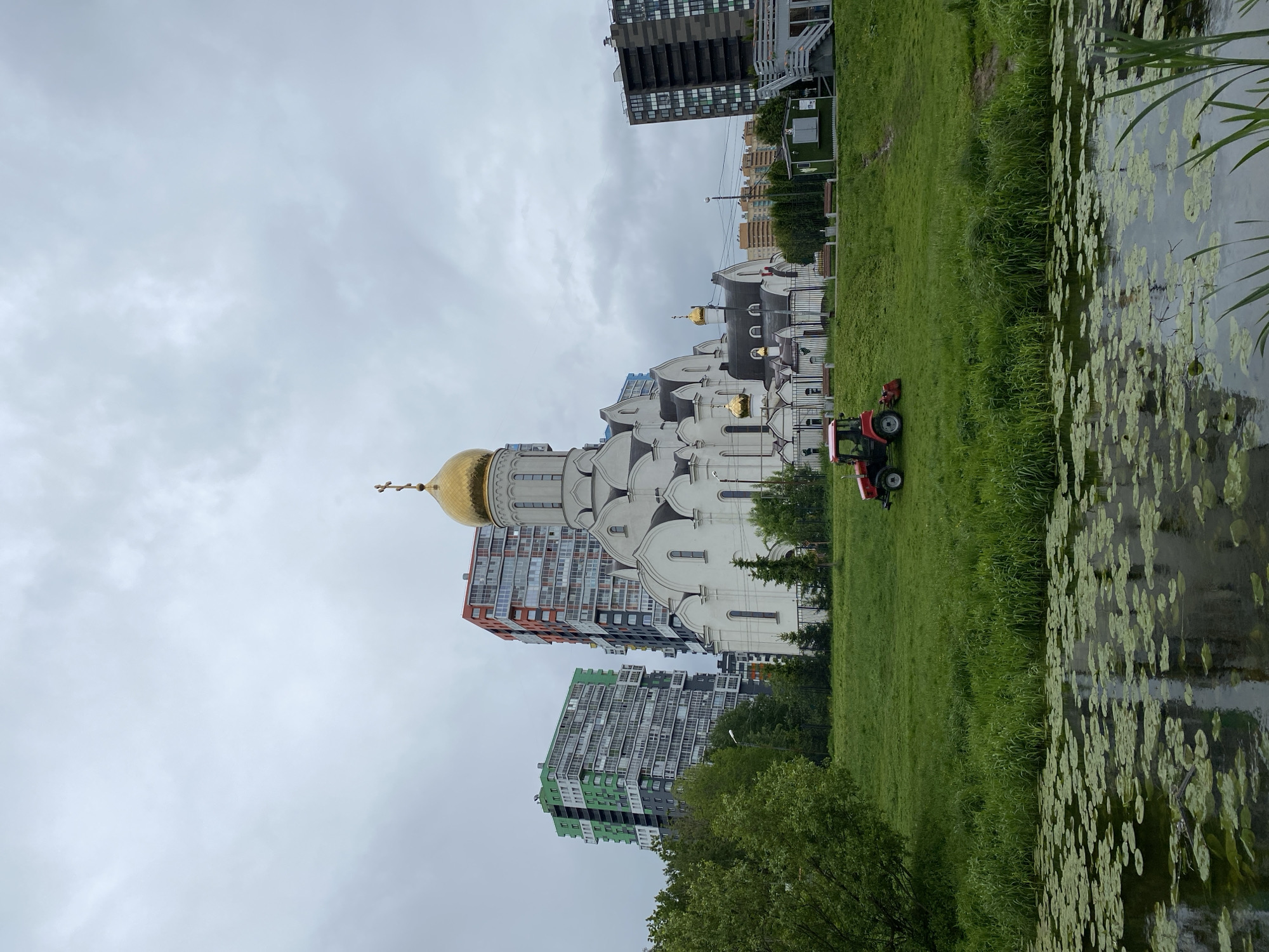 A ground-level, medium shot depicts a cityscape on an overcast day. A white cathedral with a prominent golden dome stands amidst modern residential buildings of varying heights and colors. A red riding lawn mower sits on the green lawn adjacent to a body of water covered with lily pads. The buildings are mostly high-rise apartments. The sky is uniformly gray.