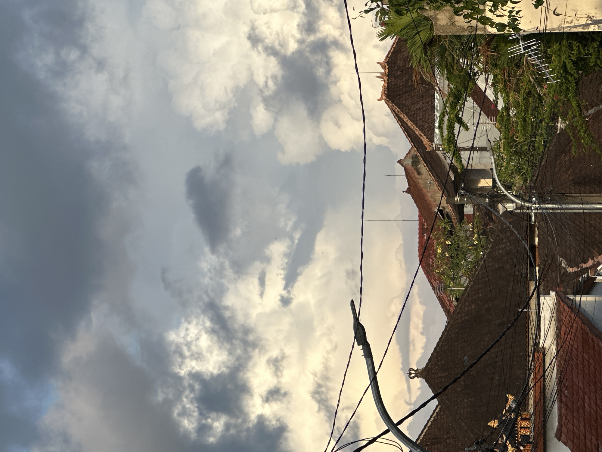 The image depicts a street scene in North Kuta, Indonesia. The sky is filled with cumulus clouds, transitioning from dark gray on the left to lighter, sunlit patches on the right. Below the sky, traditional Indonesian architecture is visible, with reddish-brown tiled roofs and ornate decorations on the rooftops. Electrical wires and a utility pole intersect the view, adding to the scene's complexity. Green foliage, including vines and palm fronds, partially obscures some of the buildings, creating a mix of natural and built environments. An antenna is visible on one of the rooftops.
