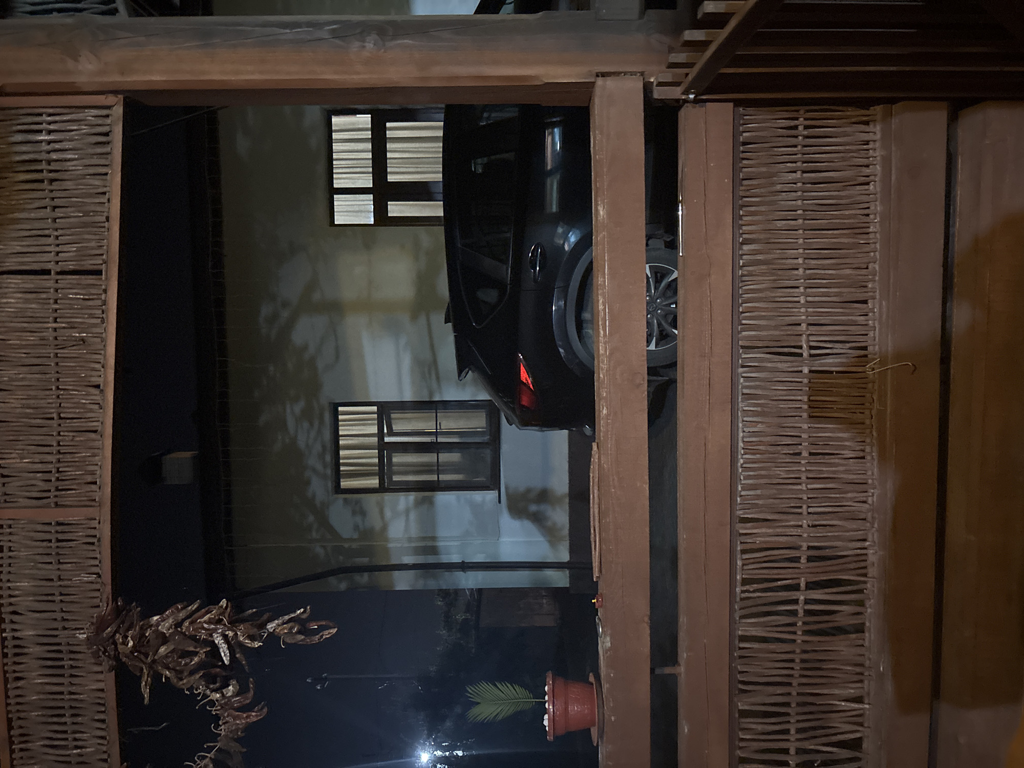 The image depicts a nighttime scene in Gudauta, Abkhazia, observed from a raised vantage point, possibly a balcony or porch constructed from dark brown wood. The foreground features a substantial wooden railing system, composed of horizontal beams and vertical elements made of narrow, irregularly arranged wooden sticks or reeds. A thick wooden support post is visible on the right.
On the left side of the wooden ledge, a small terracotta pot contains a green plant, possibly a palm sprout, with white pebbles at its base. To the right of the pot, a small orange tea light candle is present.
Behind the wooden railing, a dark-colored SUV or crossover vehicle is parked, with its rear section visible. The vehicle's left taillight is illuminated red, and portions of its wheels are visible beneath the railing.
In the background, a building with a light-toned, possibly pale blue or grey, facade is present. Two windows are visible on this building. The left window is partially open and displays a light-colored curtain. The right window shows a similar light-colored curtain, drawn closed. Shadows, consistent with tree branches or foliage, are cast upon the building's facade. The upper left area reveals a cluster of dried red peppers, strung together and hanging from an overhead structure, possibly an extension of the wooden framework. The overall environment is dimly lit, indicative of night, with internal light sources illuminating the building's windows. No individuals are visible in the scene.