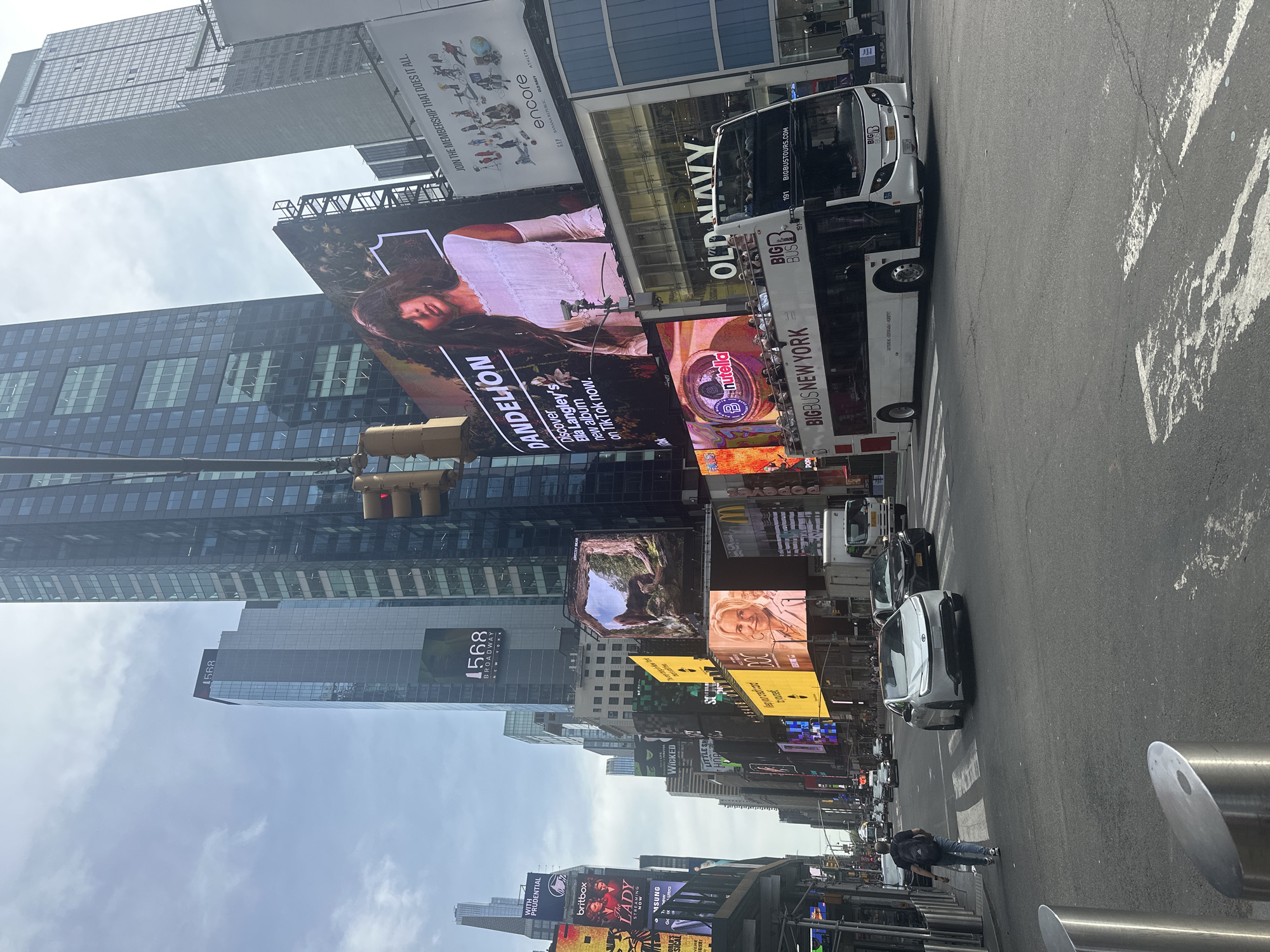 The image depicts a view of Times Square in New York City, showcasing a street scene with multiple billboards displaying advertisements. A double-decker Big Bus New York tour bus is parked on the right side of the street. Several cars are visible, including a white car. A person is walking on the sidewalk. The environment includes skyscrapers and street furniture, such as metal bollards along the sidewalk. The billboards feature a variety of advertisements for products like Nutella, Old Navy, and musicals like "Wicked". An advertisement for the album "Dandelion" by Ella Langley is also visible.