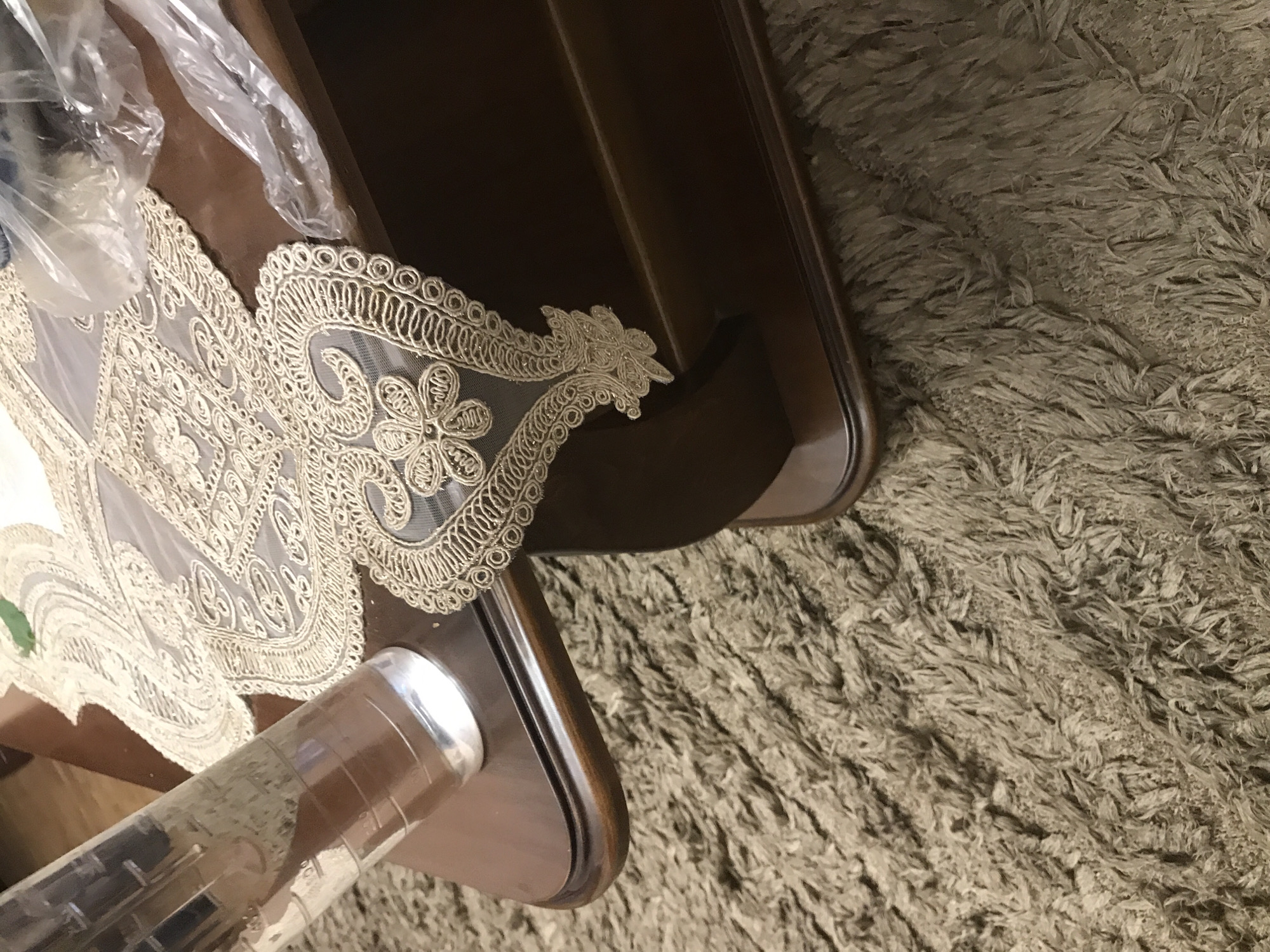 The image shows a top-down view of a dark wooden table partially covered with a light-colored, ornate doily and some plastic wrapping. A clear, cylindrical object with a silver top is visible at the bottom of the frame, resting on the table. The table is positioned on a shaggy, beige carpet. The location is not discernible from the image.