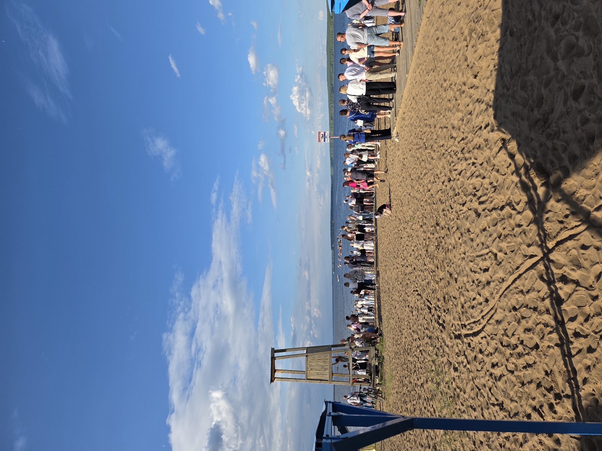 A wide shot captures a beach scene on a sunny day in Chaikovsky, Russia. A crowd of people is gathered near the water's edge, some standing and observing while others are closer to the shore. A wooden lifeguard tower stands prominently on the sand, with a bicycle leaning against its base. The sand is marked with footprints and shadows, suggesting recent activity. The water stretches out to the horizon, with boats visible in the distance and a tree-lined shore completing the background. The sky above is a clear blue with scattered white clouds.