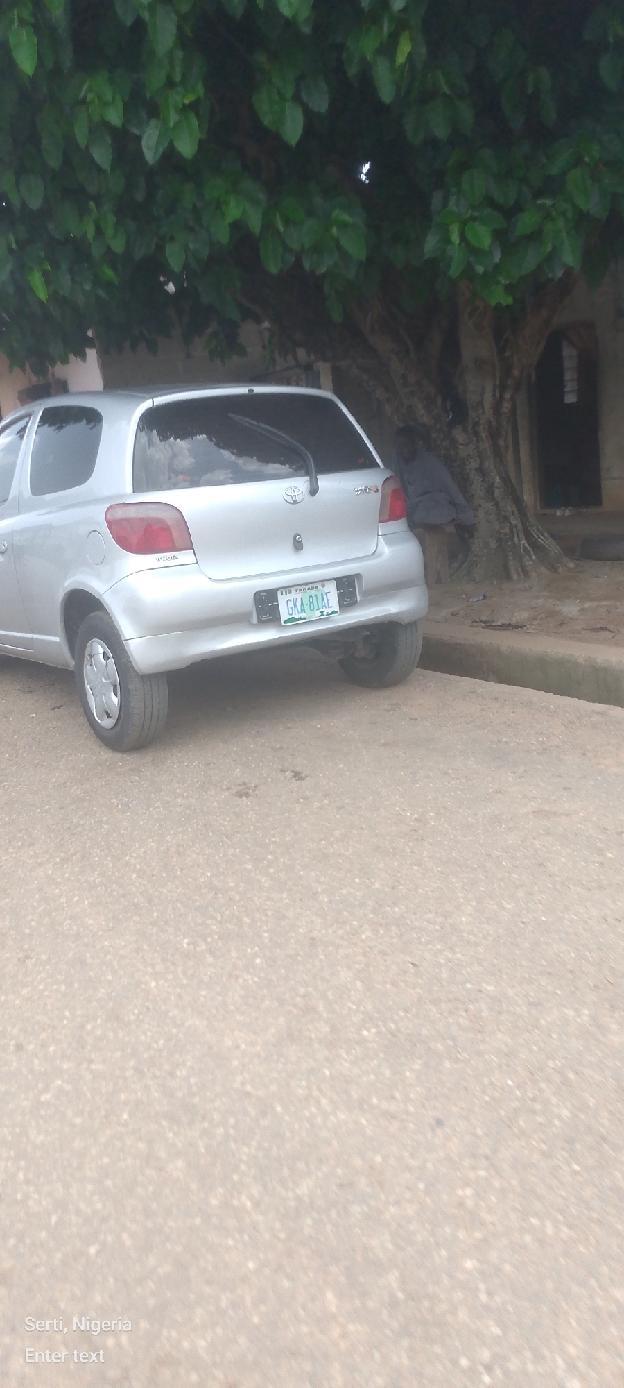A silver Toyota Vitz hatchback is parked on a dirt road under a large, leafy tree. The car's license plate reads "GKA 81AE". To the right of the car, a person is seated on a log, leaning against the tree trunk. The environment appears to be an outdoor urban setting.
