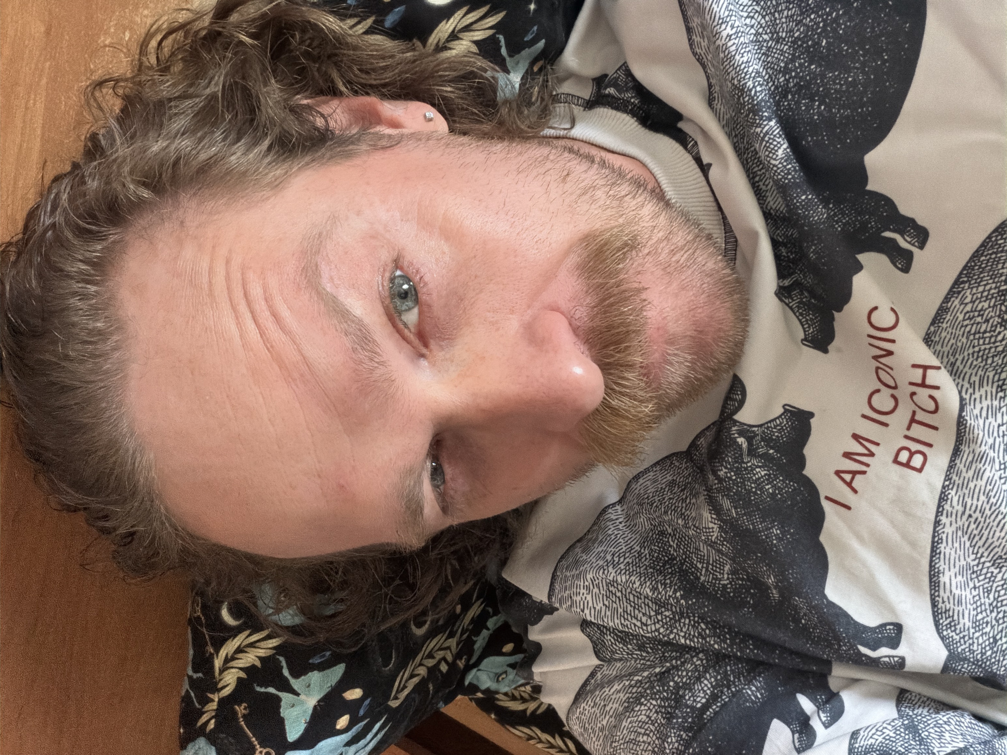 A Caucasian male with light skin, light blue eyes, and light brown hair and beard is pictured from a close-up, slightly elevated perspective. He is lying down, possibly on his back, with his head slightly tilted. He has a stud earring in his left ear. He is wearing a white shirt with a black and white design featuring what appear to be illustrated bears and the text "I AM ICONIC BITCH" in red letters. Part of a dark-colored garment with a floral pattern is visible in the lower left corner of the frame. A wooden surface, possibly a table or floor, is also visible on the left side. The photo does not provide any details to determine a location in Saint Petersburg, Russia.