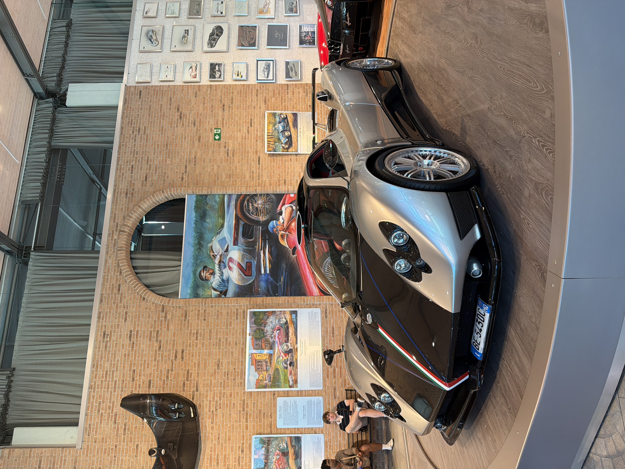 This image depicts the interior of a museum or exhibition space, likely within the city of Sant'Anna, Italy, showcasing a collection of automotive art and vehicles. The primary subject is a silver Pagani Zonda supercar, prominently displayed on a circular wooden platform. The car features a distinctive carbon fiber front with Italian flag racing stripes. In the background, a brick wall is adorned with numerous framed sketches and paintings of classic race cars and racing scenes. To the left of the supercar, a large mural depicts a vintage race car with a driver numbered '2'. Two young men are seated on a bench in the mid-ground, observing the exhibit. The environment includes exposed steel beams and ceiling structures, as well as draped curtains, suggesting an industrial or modern architectural setting. A red supercar is partially visible behind the silver Pagani.