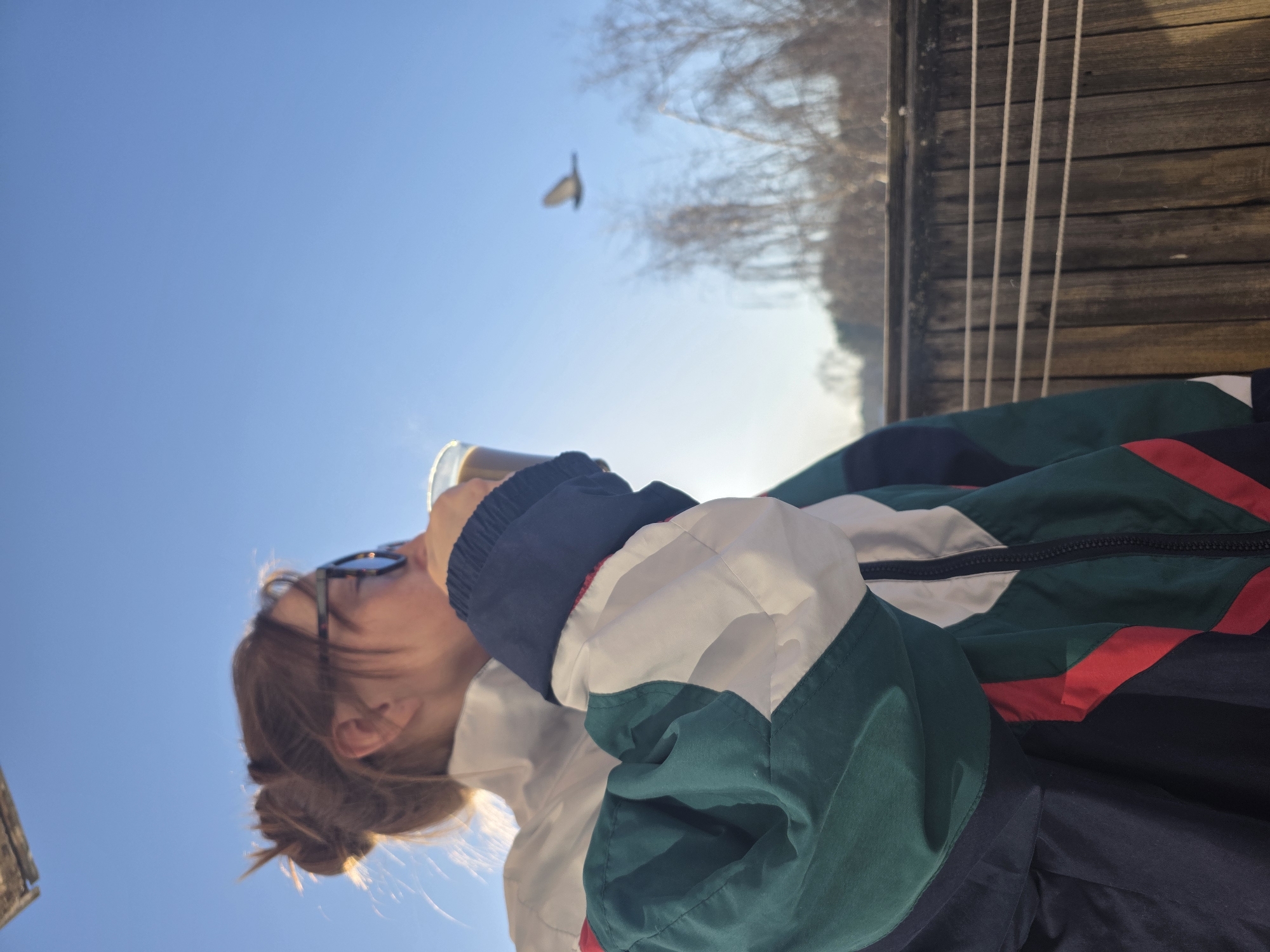 A person in a multi-colored jacket is drinking from a cup under a clear blue sky. A bird is flying in the background above some trees. The photo appears to have been taken from a low angle, looking up at the subject. In the background are what appears to be a wooden structure and more trees. There is not enough information to know if this image was taken in the city of Chaikovsky, Russia.