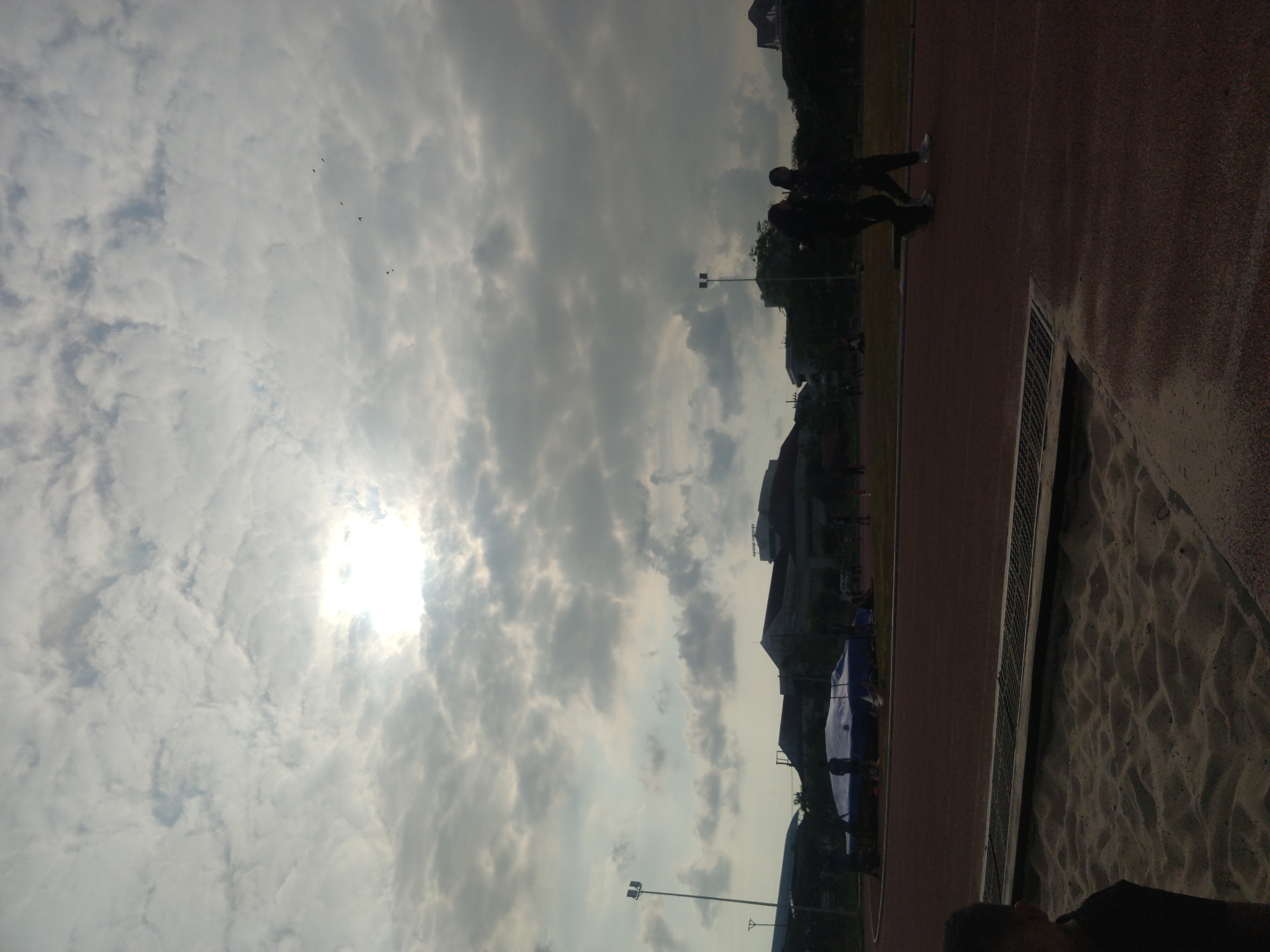 The image shows a track and field event, likely a long jump competition. In the foreground, a sand pit is visible, with a track and stadium in the background. Two individuals are walking on the track in the middle ground, and several other people are scattered around the stadium. The sky is cloudy, with the sun peeking through. The scene appears to be taking place in Surabaya, Indonesia, based on the architectural style of the buildings in the background and the presence of lush greenery, which is typical of the region. The overall atmosphere is one of a sporting event taking place on a slightly overcast day.