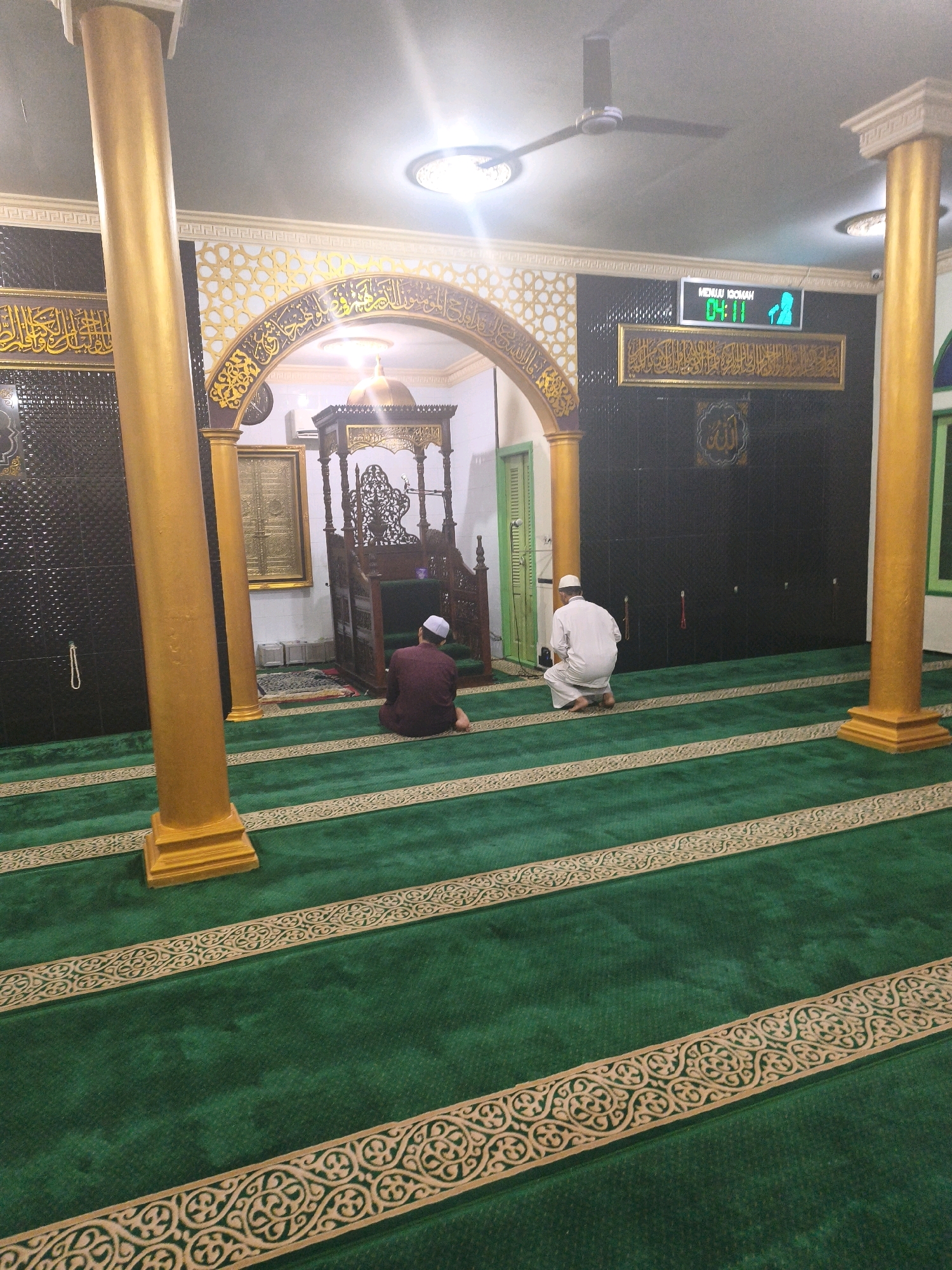 The image shows the interior of a mosque in Samarinda, Indonesia, with green carpets featuring decorative golden borders. Two men are visible, both wearing traditional Islamic attire. One man, wearing a maroon robe and white cap, is kneeling on the carpet facing away from the camera, towards the mihrab (prayer niche). Another man, dressed in white, is also kneeling on the carpet, facing the same direction. A wooden minbar (pulpit) is positioned to the left of the frame, near an arched doorway. Above the prayer area, an electronic display shows "MENUJU IQOMAH" and "04:11", indicating prayer time. The mosque features golden pillars and intricate Arabic calligraphy on the walls. A ceiling fan is also visible.