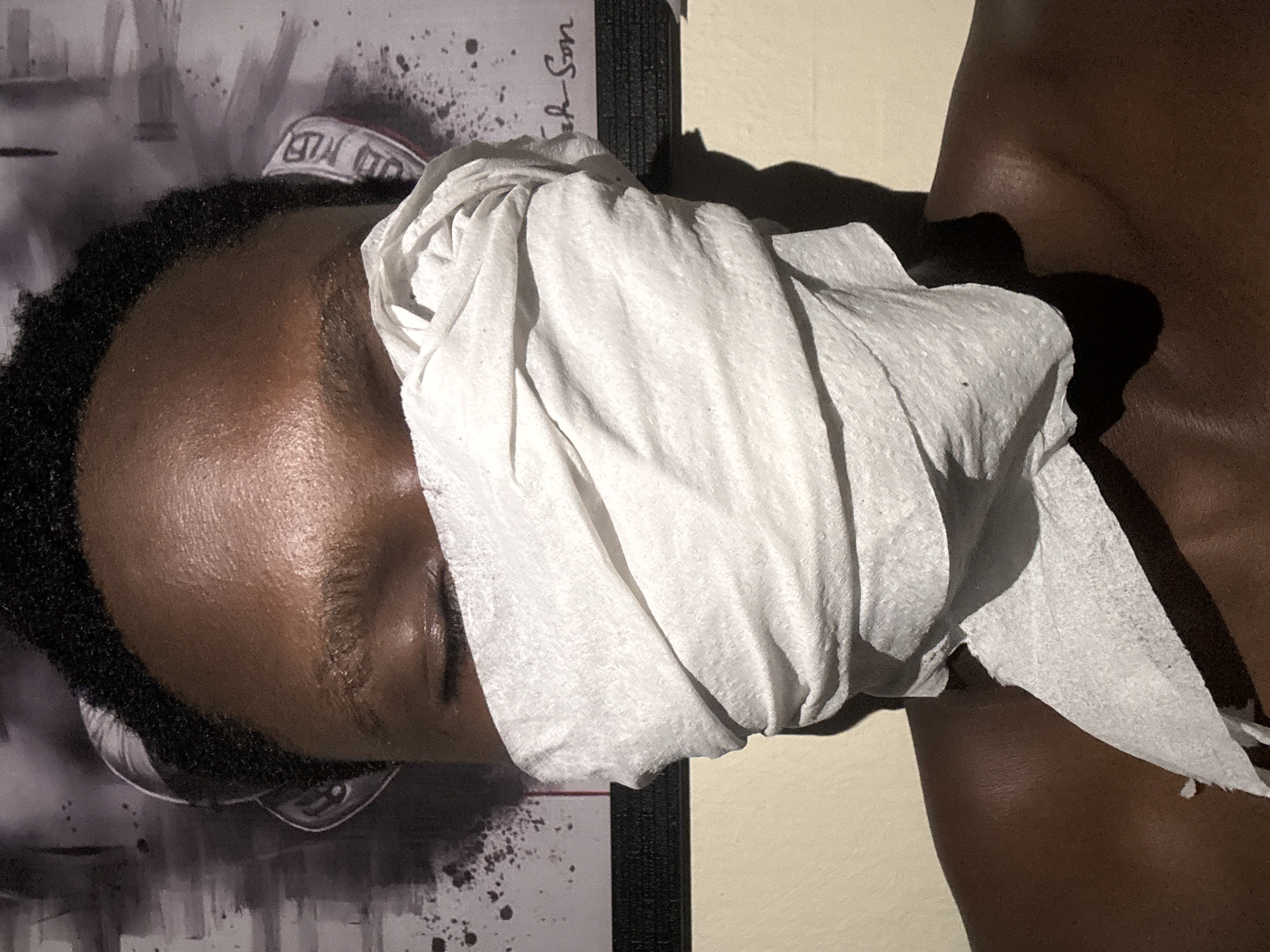 The scene is located in the city of Ihiagwa, Nigeria. The image depicts a close-up portrait of a person from the forehead to the upper chest, indoors. The individual has dark skin and short, tightly curled dark hair. Their eyes are closed. A substantial amount of white, textured tissue paper is wrapped around the lower half of their face, completely obscuring the nose, mouth, and chin, forming multiple folds and layers. A loose piece of tissue hangs down towards the left shoulder. The person's shoulders and upper chest are bare. In the background, on both sides of the head, a light-colored headband or head covering with dark markings is partially visible beneath the hair. Behind the individual, a framed poster or artwork is visible, featuring a white or light grey background with dark, abstract splatter patterns. Partial text, including "eh Son", is discernible on the right side of the poster. A dark frame border runs below the artwork. The lower background consists of a plain, light beige wall. Strong lighting illuminates the scene, casting distinct shadows on the tissue paper and the person's neck.