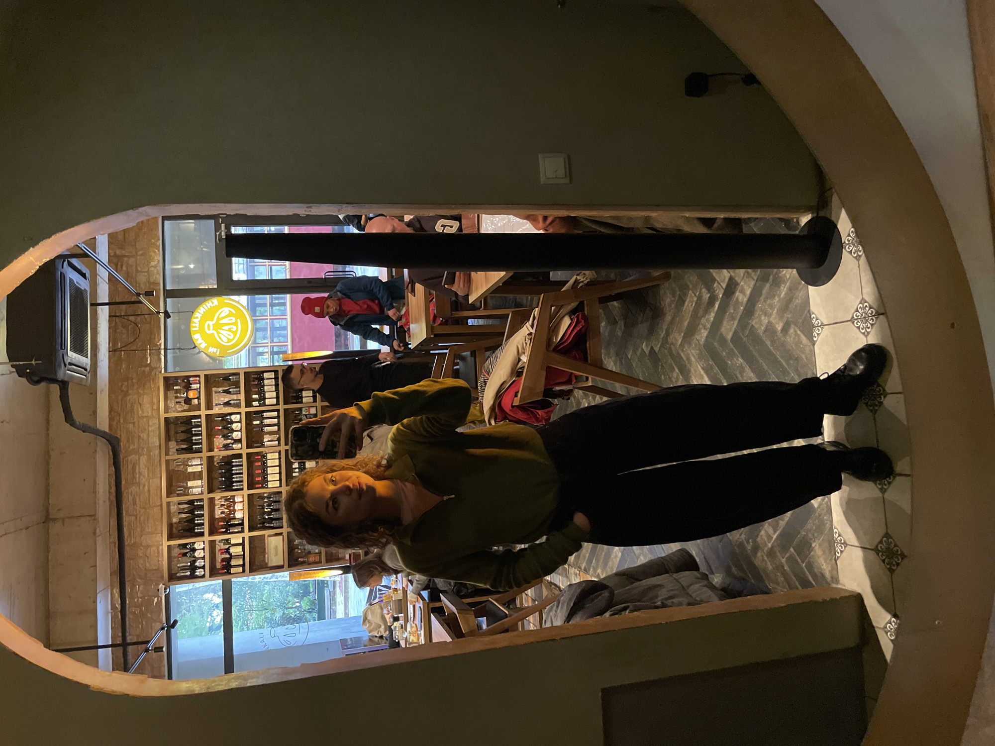 A woman stands in front of a large mirror inside a restaurant. The mirror reflects the restaurant interior. The woman is taking a selfie, dressed in black pants and an olive-green sweater. The restaurant has wine bottles displayed in a shelf in the background. Tables and people are visible in the reflection. A neon sign is also visible in the reflection. A pole is near the mirror. The location is Tbilisi, Georgia.