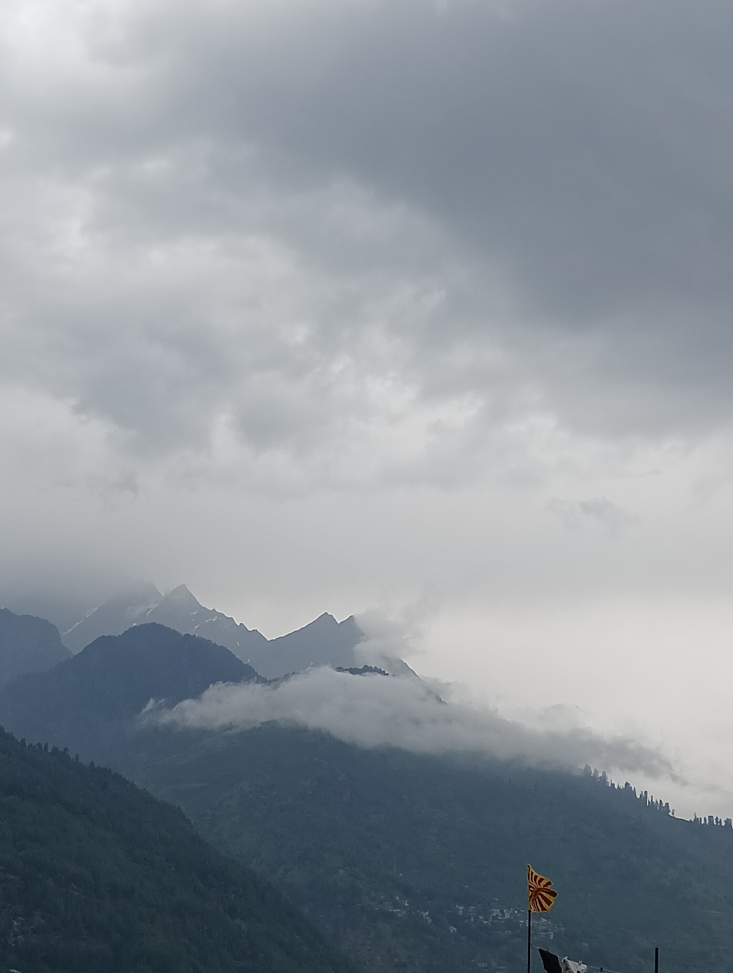 A dramatic, overcast sky looms over a mountainous landscape, likely Vashisht, India. The sky is filled with dense, gray clouds, suggesting a potentially rainy or foggy day. Below the clouds, jagged mountain peaks emerge, some dusted with snow or shrouded in mist. The lower slopes of the mountains are covered in dark green vegetation, appearing dense and forested, with hints of small settlements nestled in the valleys.
In the foreground, a solitary flag stands on a pole. The flag is a striking visual element, featuring a pattern of alternating red and yellow stripes radiating from a central point, resembling a sunburst or a spinning wheel motif. The flag appears to be fluttering slightly. The overall mood of the image is somber and majestic, emphasizing the raw beauty of the mountain environment under a heavy sky. The time of day is not clearly discernible due to the overcast conditions, but the diffused light suggests it is not midday. No visible text is present in the image.
