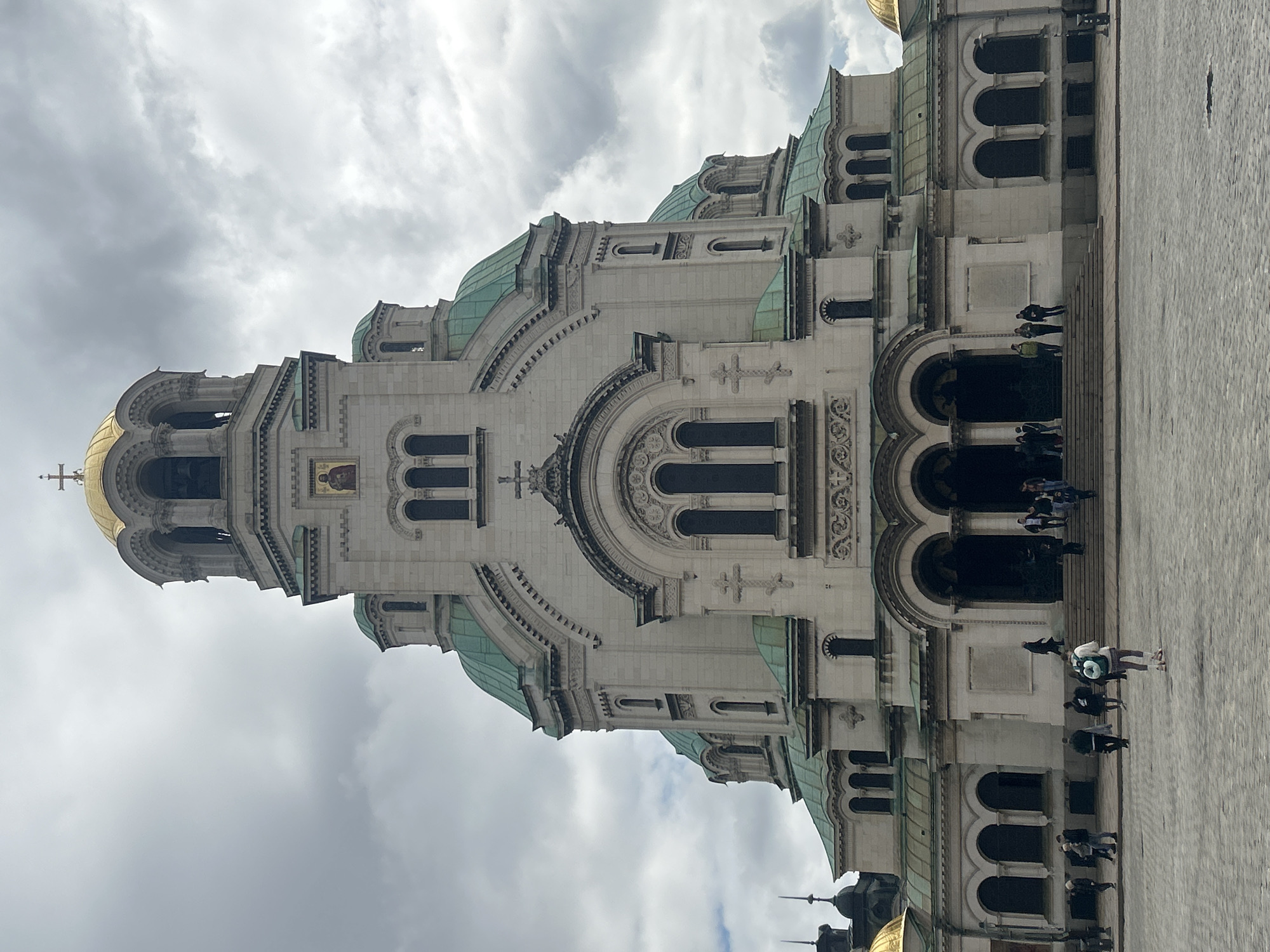 The image shows the Alexander Nevsky Cathedral in Sofia, Bulgaria, angled so that the sky is to the left of the frame. The cathedral is made of light-colored stone and has multiple domes with green roofs and a golden bell tower. People can be seen walking near the entrance of the cathedral on the right side of the image. The sky is overcast with grey clouds.