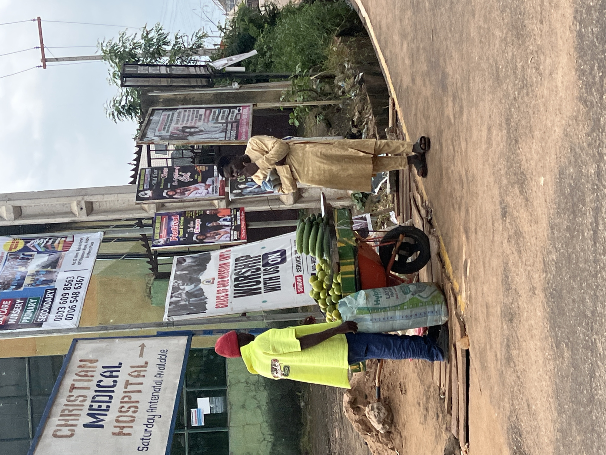 In Okpanam, Nigeria, outside a building with signs for "CHRISTIAN MEDICAL HOSPITAL" and various advertisements, a street vendor is seen pushing a wheelbarrow laden with produce, predominantly cucumbers and what appear to be oranges or similar citrus fruits. Beside the wheelbarrow, a man wearing a bright yellow long-sleeved shirt and a red beanie stands, holding a large white bag with "Hybred" visible on it. To the right of the vendor, another man in a long, tan traditional outfit stands with his arms crossed, holding a light blue cloth. The ground is paved with a mixture of asphalt and dirt, with several wooden planks laid out. The background shows dense green foliage and a cloudy sky.
