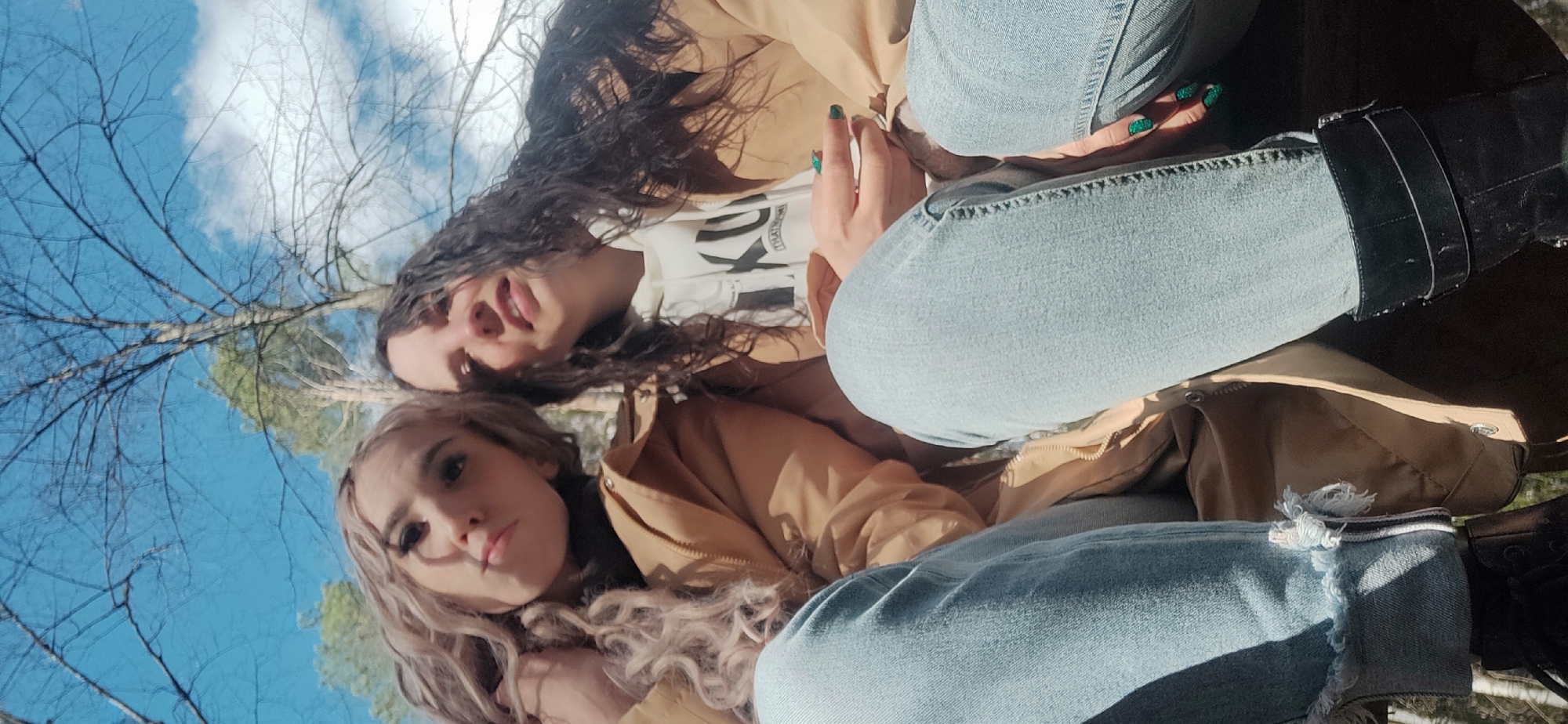 Two young women pose for a selfie outdoors. One woman has long, dark curly hair, wearing a white t-shirt, a tan jacket, and light blue jeans. The other woman has long, blonde curly hair, wearing a tan jacket and light blue jeans. The sky is visible in the background, with bare tree branches against a blue sky. The location is Unknown, Unknown.