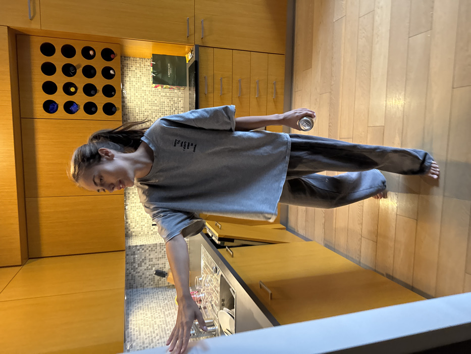 The image depicts the interior of a contemporary residential kitchen. While the request specifies Warsaw, Poland, no identifying features within the image allow for the precise pinpointing of a specific address or neighborhood within the city.
A single individual, a young adult female, is captured slightly left of the image's center, facing towards the right with her head slightly tilted upwards. She has light brown hair tied in a ponytail and is dressed in a grey oversized short-sleeved t-shirt, which features black text on the left chest reading "sorry i'm late, i didn't want to come." She is also wearing grey sweatpants and is barefoot. Her right arm is extended horizontally forward, with her hand appearing to touch or reach towards a white vertical surface on the far left of the frame. In her left hand, she holds a silver-colored beverage can.
The kitchen features light orange-toned wooden cabinets, both upper and lower, complemented by a dark grey countertop. A light-colored mosaic tile backsplash is visible above the counter. On the left, a stainless steel sink is present, next to which sits a dish drying rack holding several clear glasses and white plates. A knife block with knives is positioned further back on the counter. An integrated wooden wine rack, containing multiple circular compartments with some bottles visible, is built into the upper cabinets on the right side of the frame. Further along the counter to the right, a dark green shopping bag bearing the name "LAUDERBACH" is visible, adjacent to a dark-colored cooktop. The floor consists of light-toned wooden planks. Undercabinet lighting illuminates the backsplash area.
