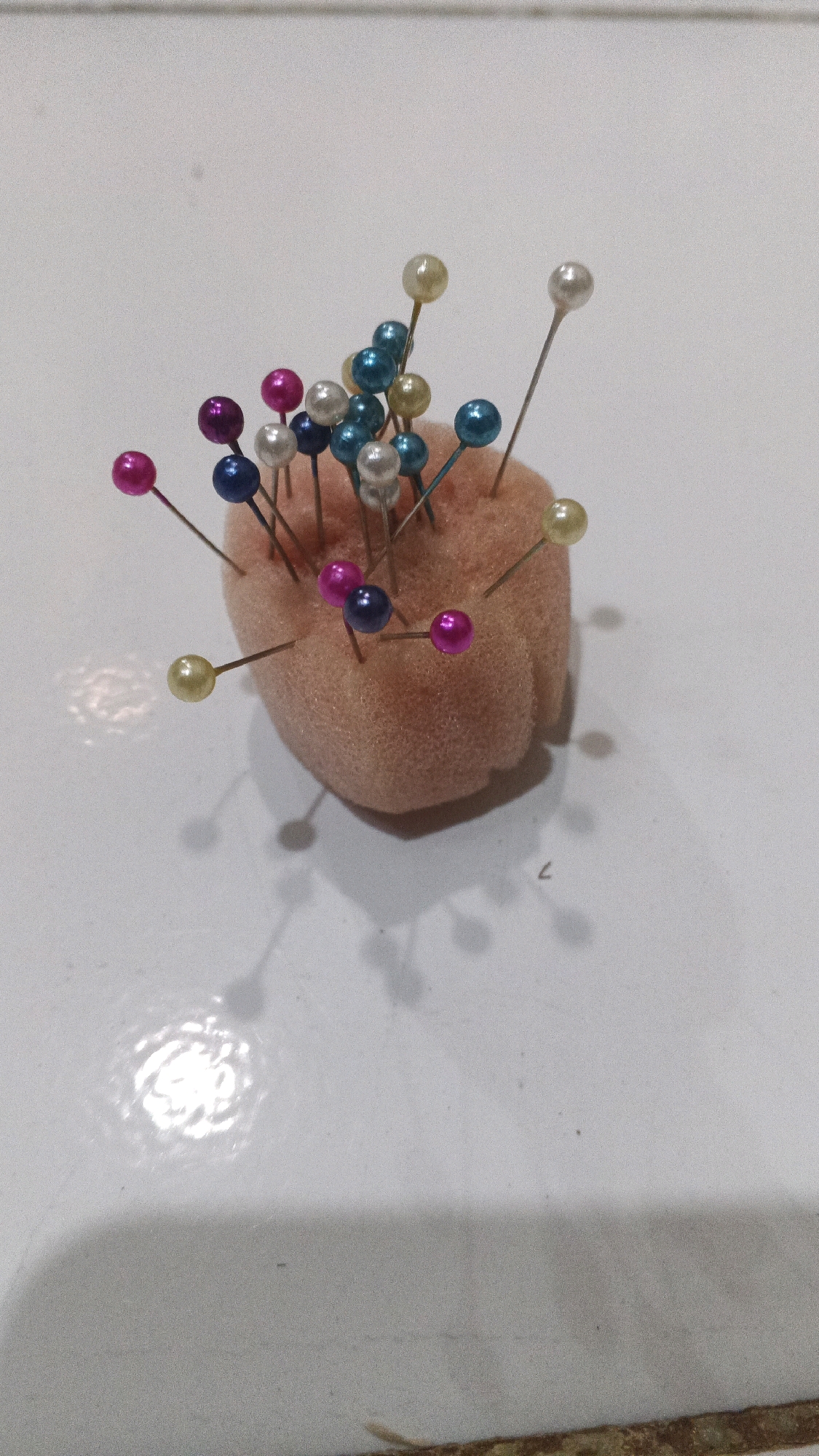 A close-up, overhead shot shows a pincushion with colorful pearl-headed pins stuck into it. The pincushion is a pale pink, round, and appears to be made of foam. The pins have heads of various colors, including pearl white, light yellow, dark blue, teal, and magenta. Some pins are oriented vertically, while others are at various angles. The background is a white surface with a subtle texture, possibly a table or countertop. The lighting creates soft shadows of the pins and pincushion on the surface. The image is sharp and clear, with a shallow depth of field that blurs the background slightly. There is no indication of any specific location in Winong, Indonesia.