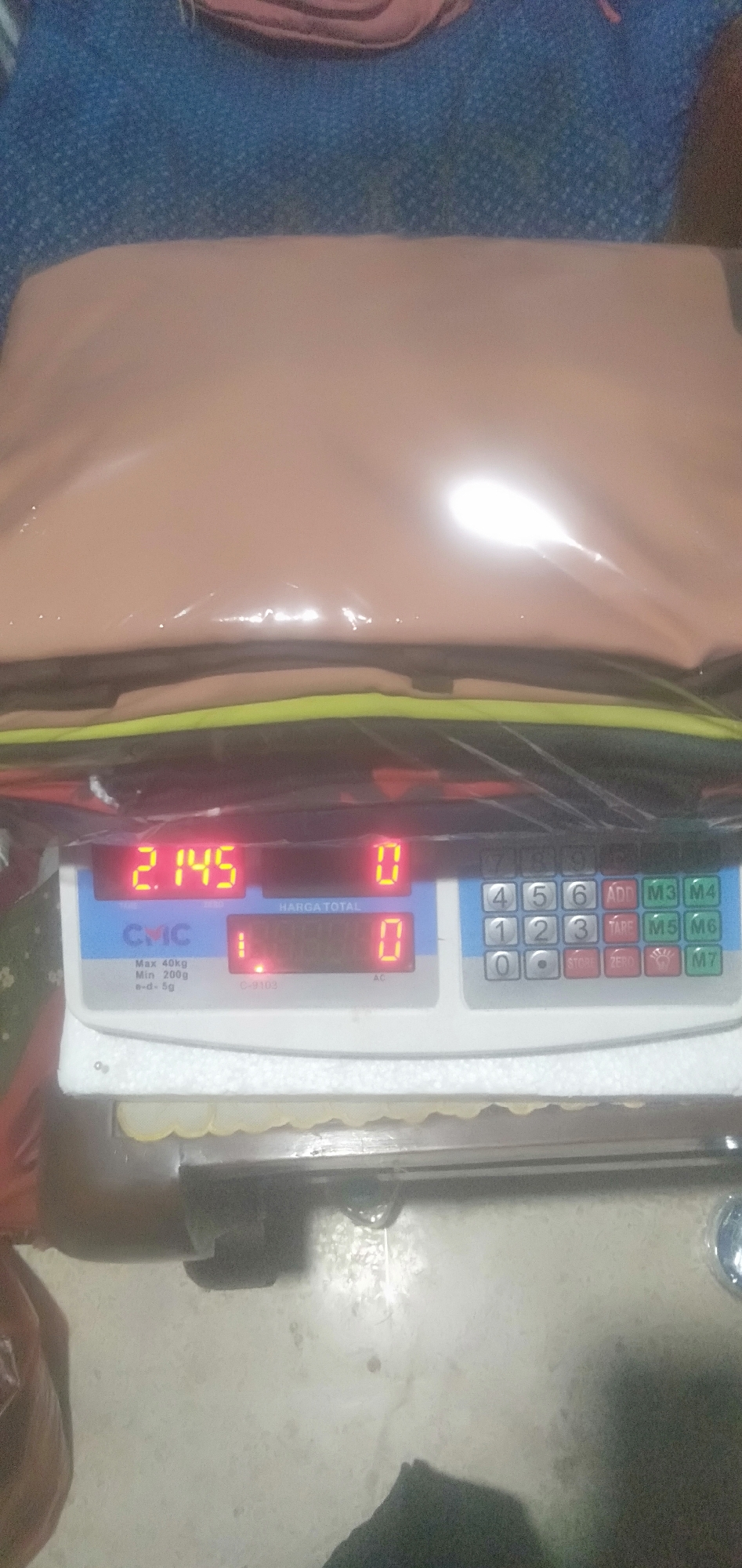 An overhead shot shows a digital scale weighing a beige-colored bag. The scale displays "2.145" on its left screen and "0" on its right screen. Below these, the scale shows "1" and "0" in red digits. The brand "CMC" is visible on the scale. The scale itself is on a speckled, light-colored floor, and a blue and red patterned fabric is visible in the background. The image is taken indoors.